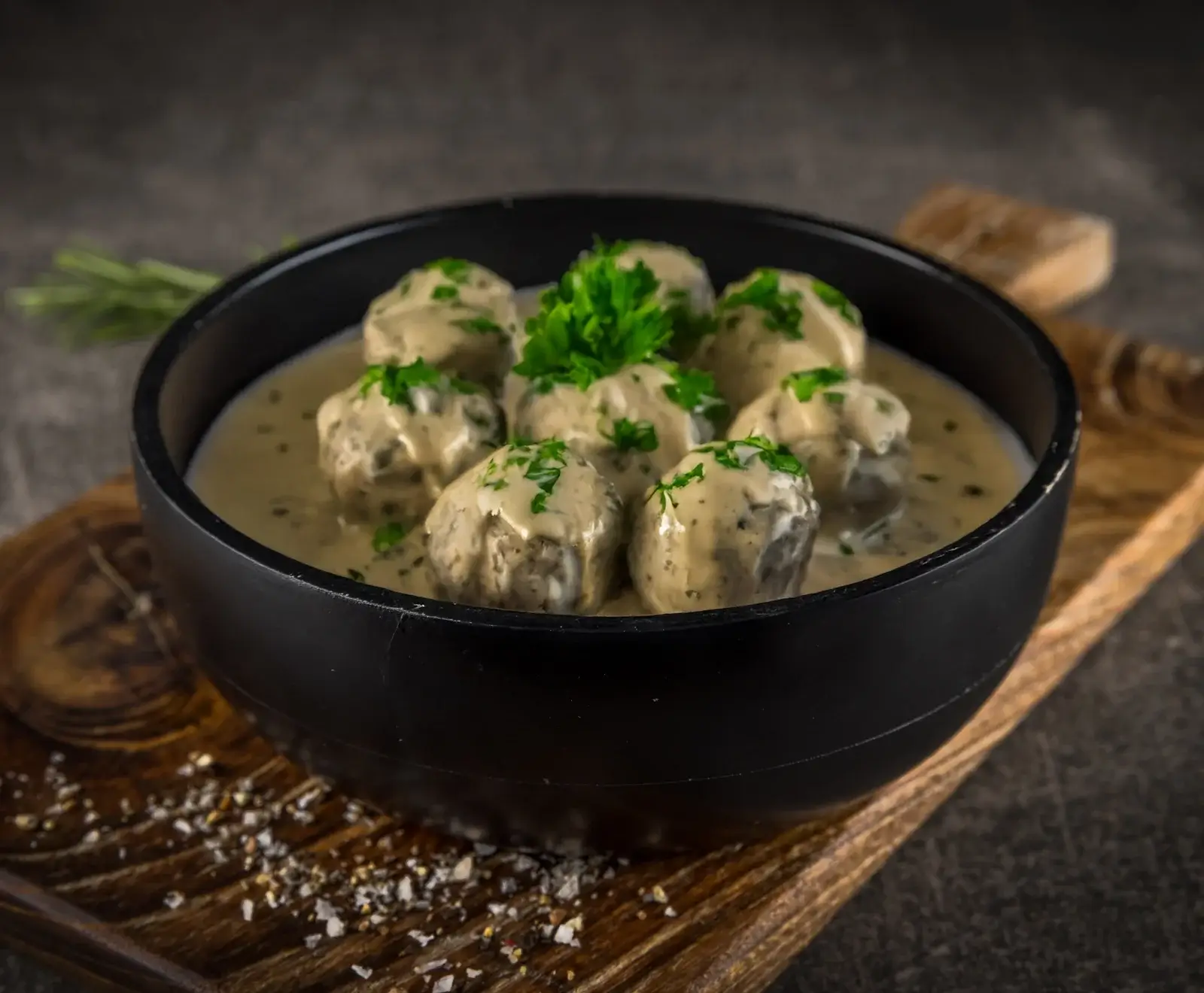 Schwarze Schale mit Fleischbällchen in cremiger Soße, garniert mit Petersilie, auf einem Holzbrett mit verstreuten Gewürzen.