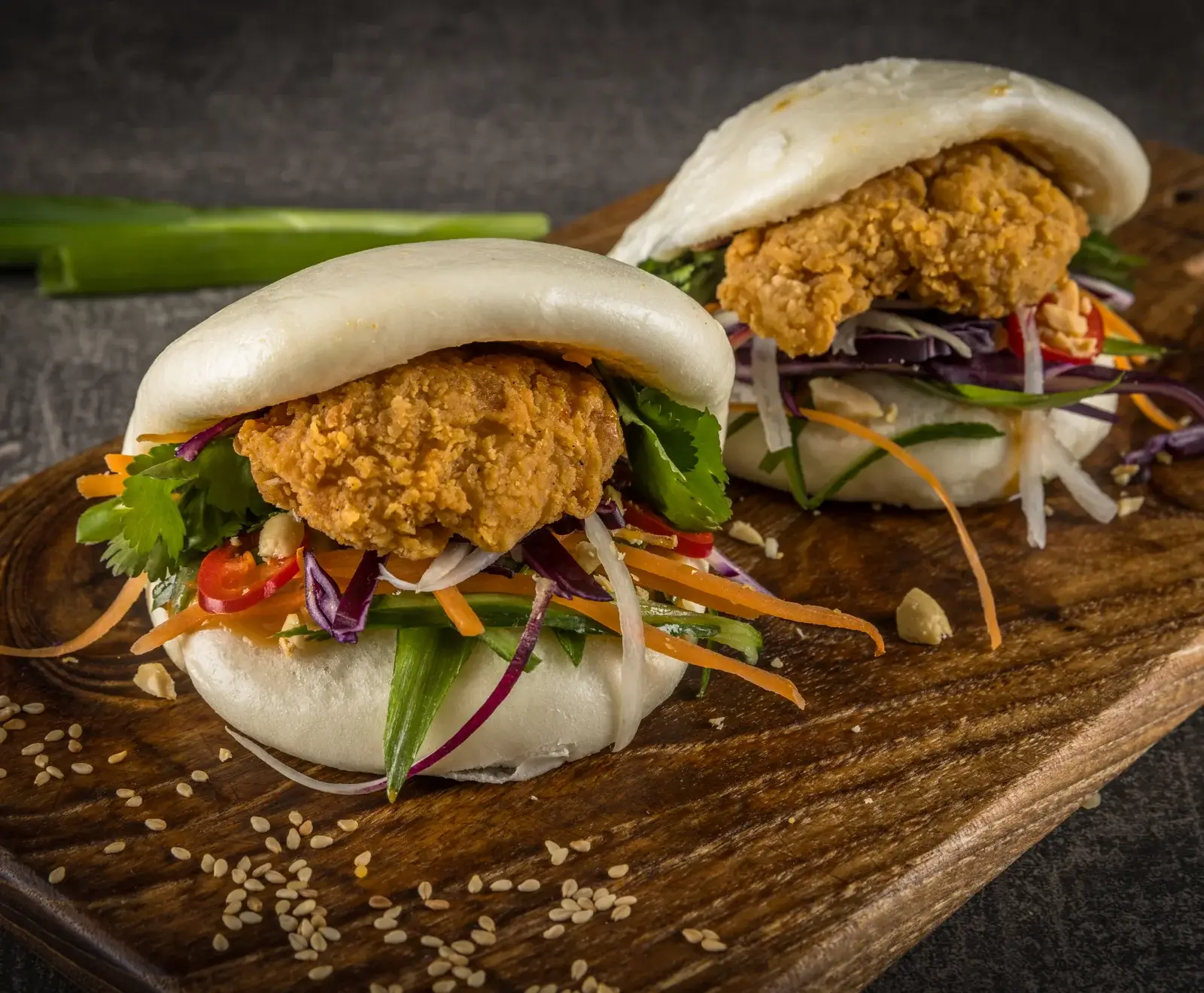 Zwei gefüllte Bao Buns mit knusprigem Brathähnchen, frischem Gemüse wie Karotten, Rotkohl, Gurken und Koriander, serviert auf einem Holzbrett mit Sesamsamen.