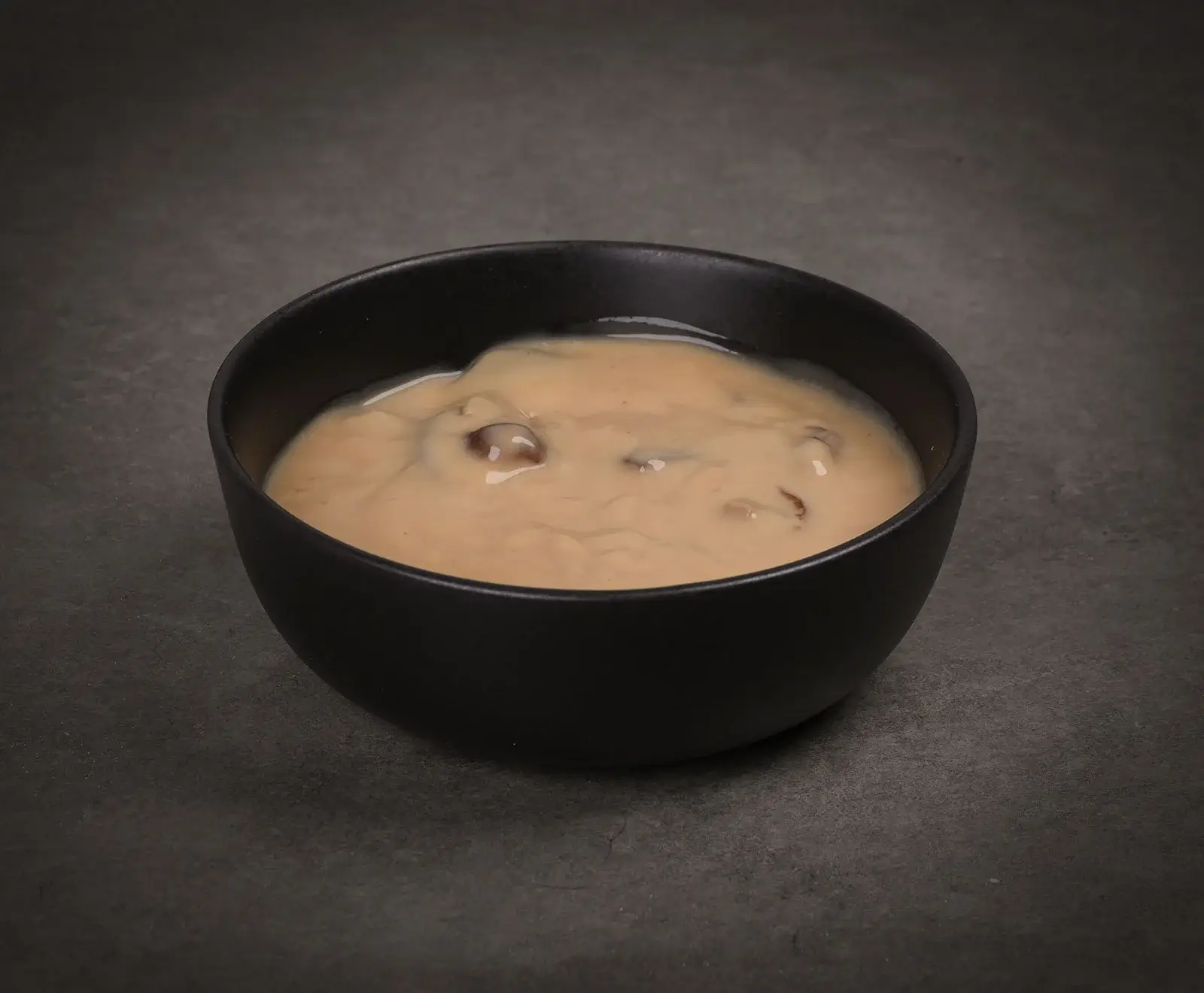 Eine schwarze Schale gefüllt mit cremiger, hellbrauner Pilzsauce, die auf einer dunklen, strukturierten Oberfläche steht. Die Sauce enthält sichtbare Pilzstücke.