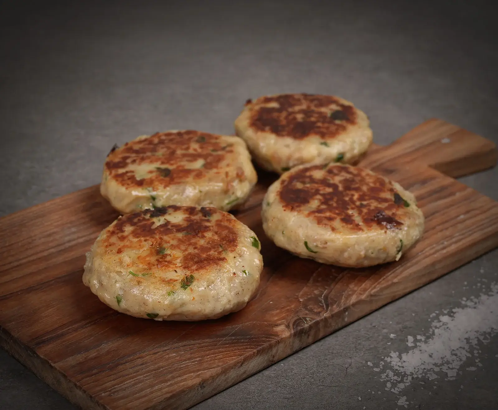 Vier goldbraun gebratene Frikadellen mit Kräutern liegen auf einem rustikalen Holzbrett. Im Hintergrund ist eine dunkle Oberfläche mit grobem Salz zu sehen.