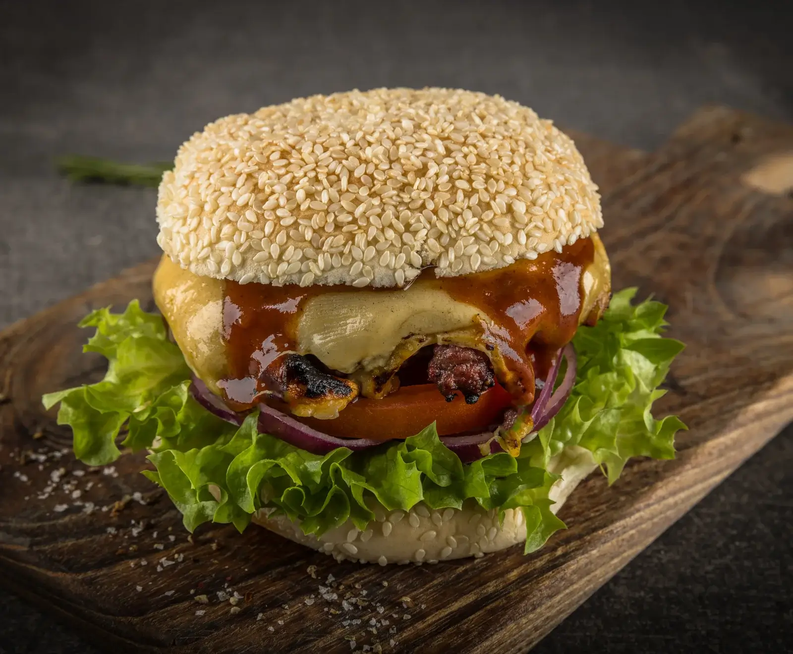Ein appetitlicher Burger mit Sesambrötchen, geschmolzenem Käse, BBQ-Sauce, gegrilltem Patty, roten Zwiebeln, Tomate und frischem Salat, serviert auf einem rustikalen Holzbrett.