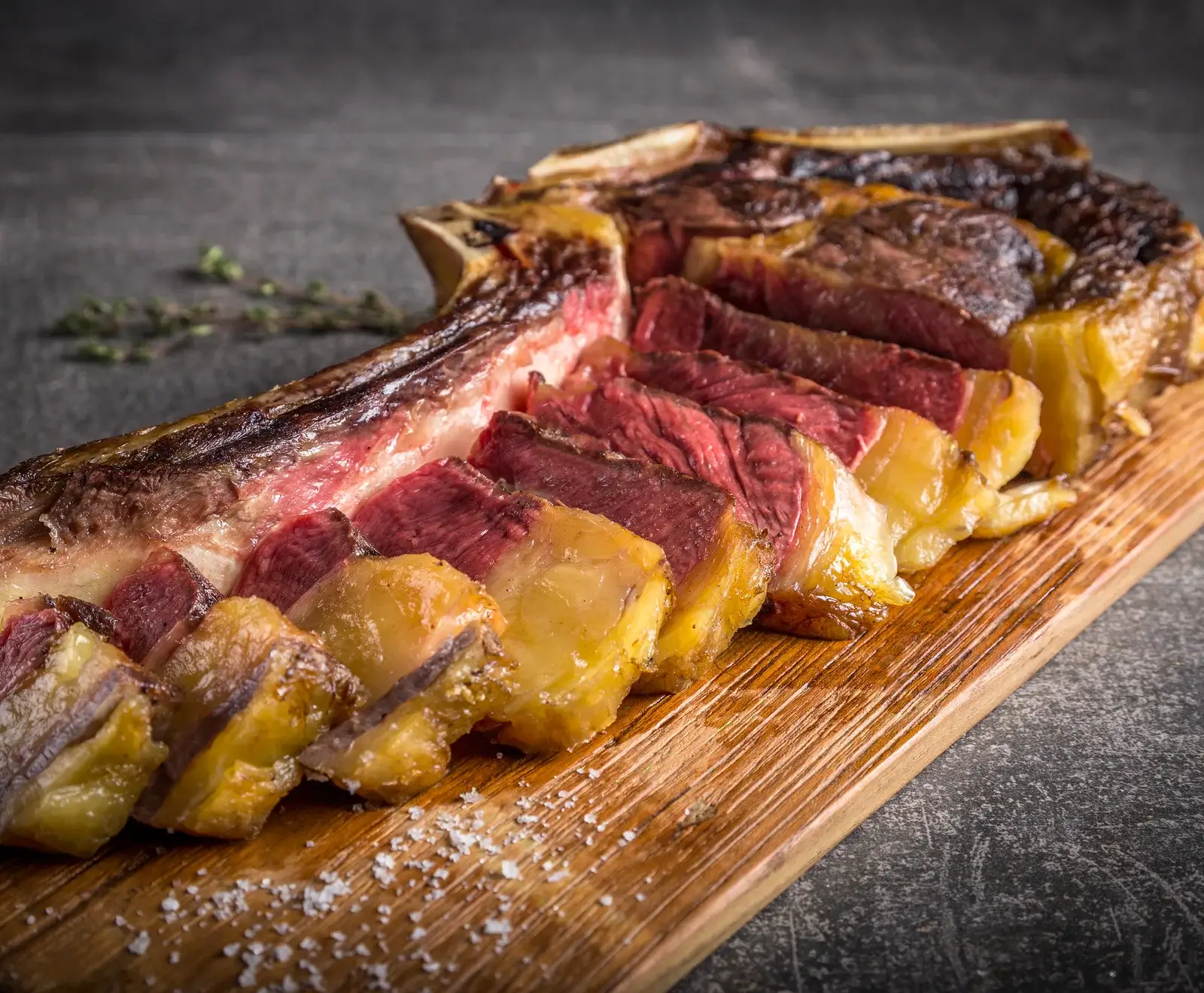 Ein perfekt gegartes, in Scheiben geschnittenes Tomahawk-Steak mit rosa Kern und knuspriger Kruste, angerichtet auf einem Holzschneidebrett mit grobem Salz. Im Hintergrund sind Kräuter zu sehen.