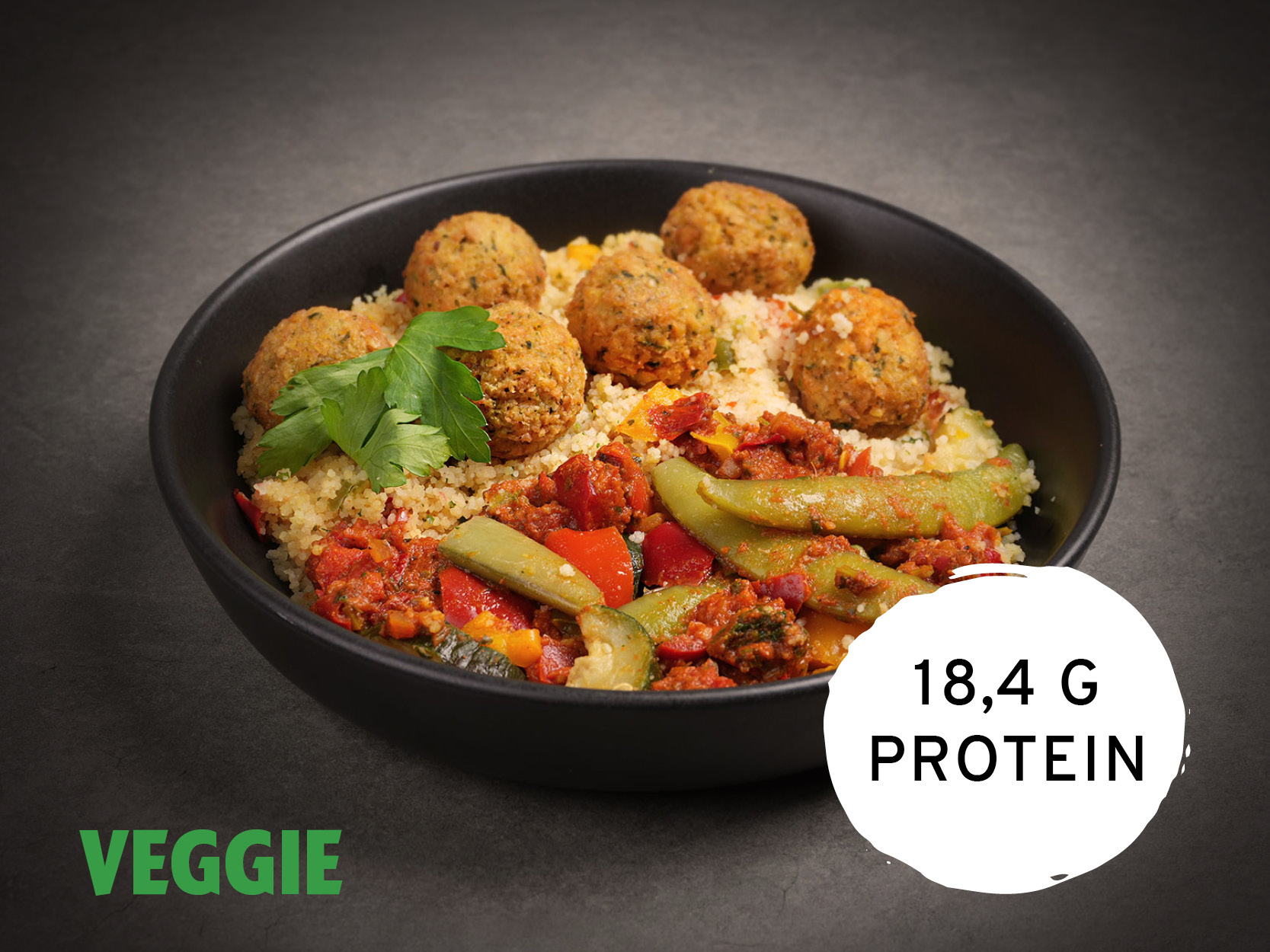 Eine schwarze Schale mit Couscous, Falafelbällchen und mediterranem Gemüse in Tomatensauce, garniert mit Petersilie. Unten links steht "VEGGIE", unten rechts "18,4 G PROTEIN".
