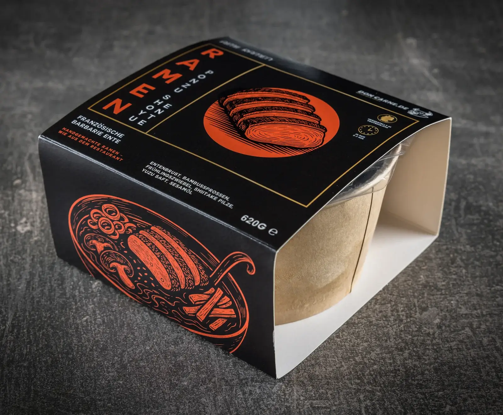 Schwarze Verpackung für Ramen mit französischer Barbarie-Ente, orangefarbene Illustrationen von Entenbrust und einer Ramenschale, Inhalt 620g, auf dunklem Untergrund.