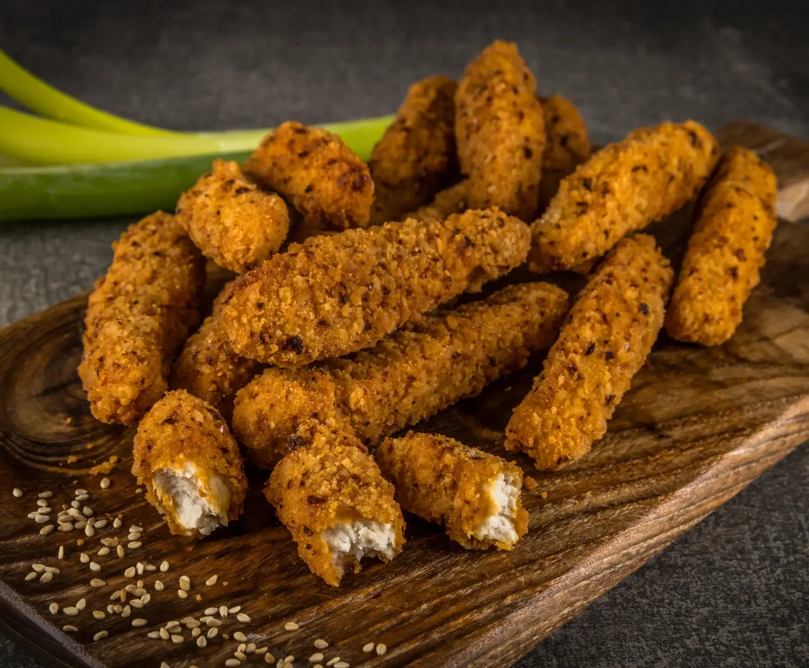Goldbraune, knusprige Hähnchen-Nuggets oder -Strips auf einem rustikalen Holzbrett. Einige sind aufgebrochen und zeigen das weiße Fleisch. Verstreute Sesamsamen und unscharfe grüne Zwiebeln im Hintergrund.