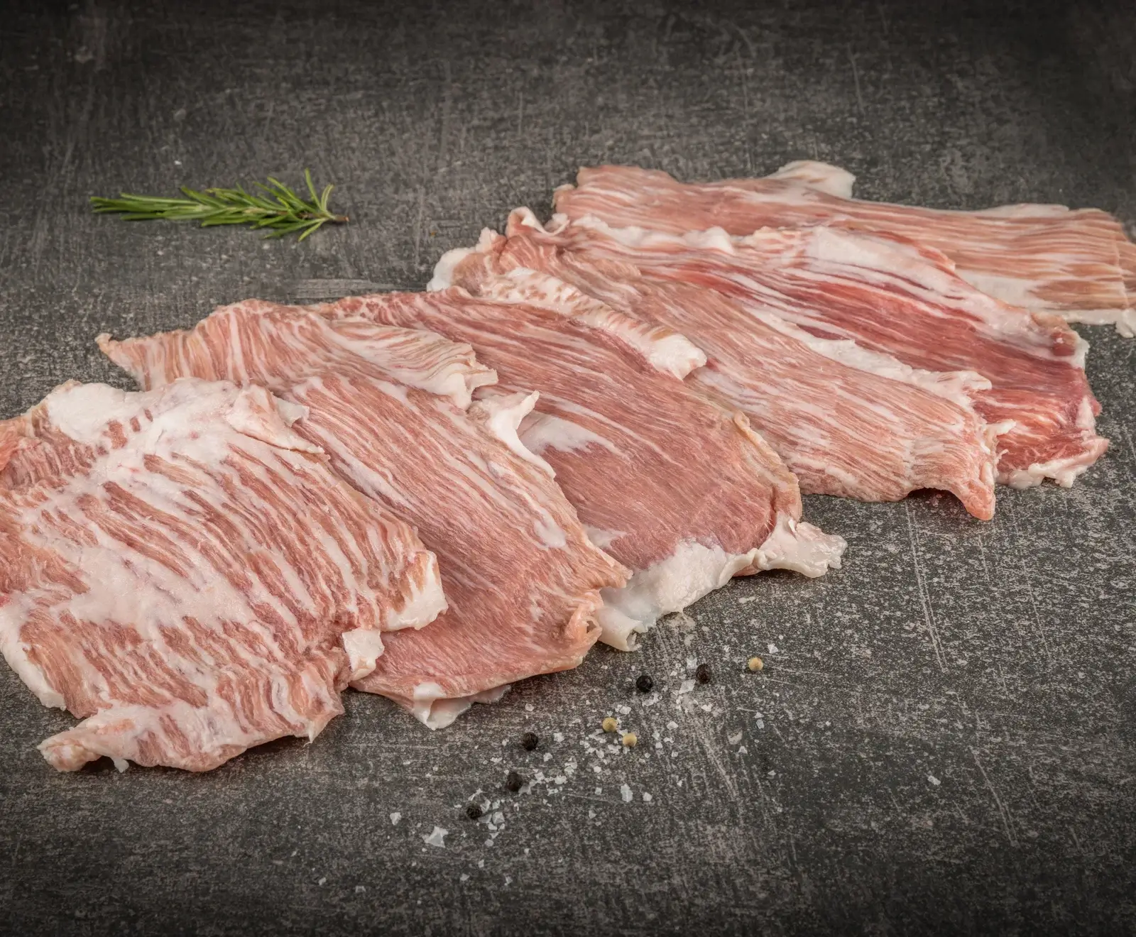 Dünne, stark marmorierte Scheiben rohes Fleisch, diagonal auf einem dunklen, strukturierten Untergrund angeordnet, mit einem Rosmarinzweig, Salz und Pfefferkörnern.