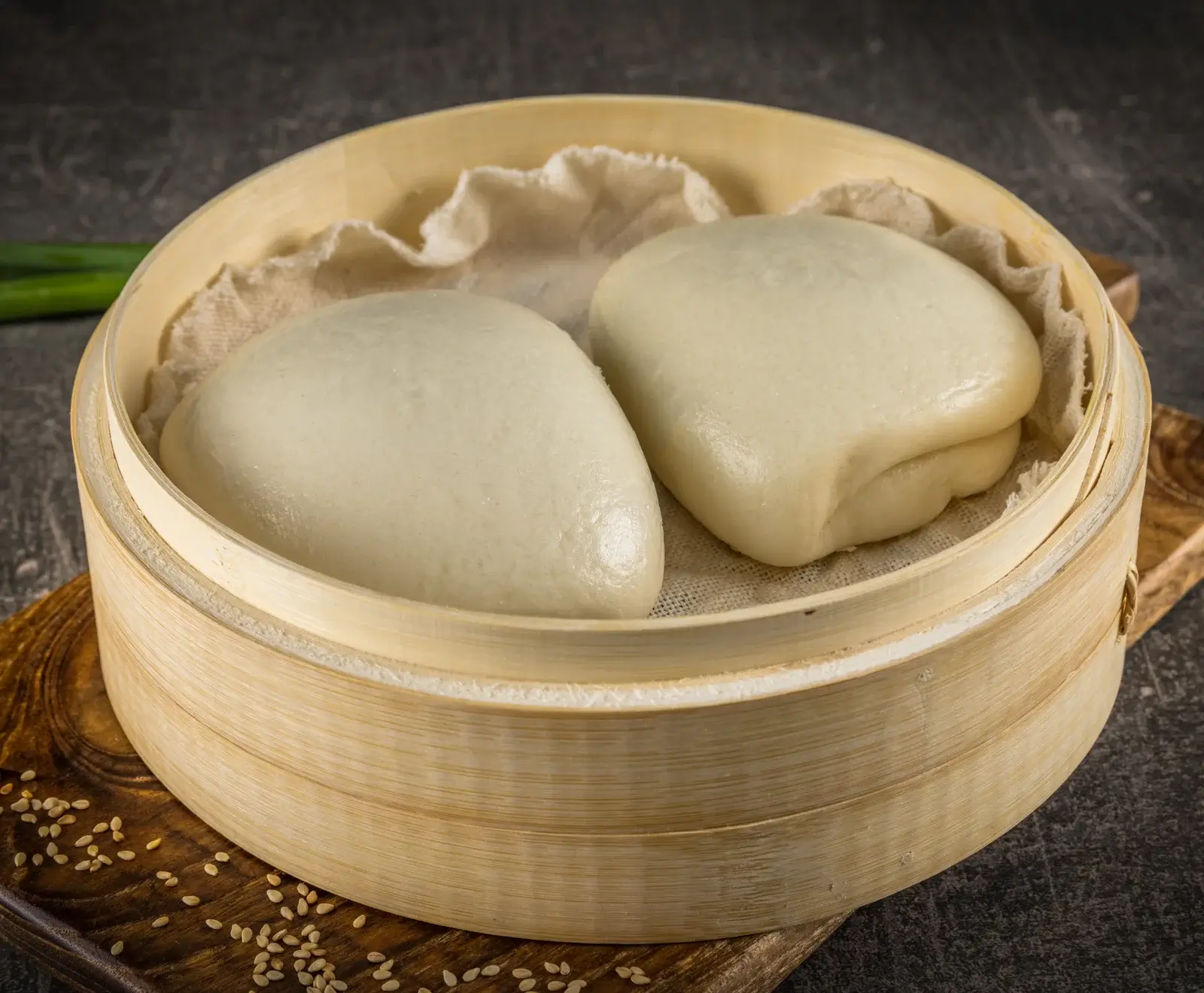 Zwei helle, gedämpfte Baozi-Brötchen liegen in einem runden Bambusdämpfer auf einem weißen Tuch. Der Dämpfer steht auf einem Holzbrett mit Sesamsamen.