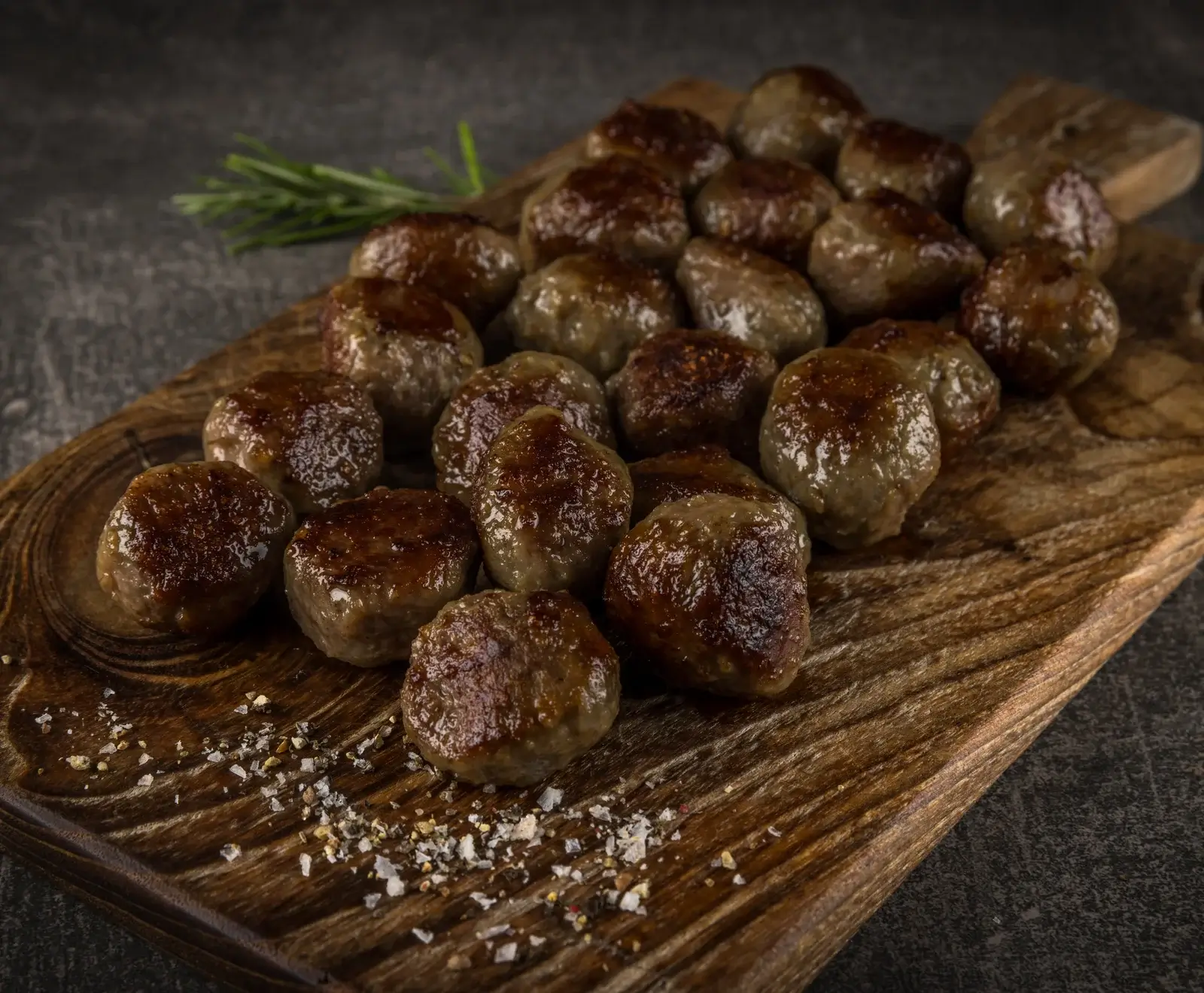 Nahaufnahme von vielen goldbraun gebratenen Fleischbällchen auf einem rustikalen Holzbrett, garniert mit Rosmarin und grobem Salz/Pfeffer.