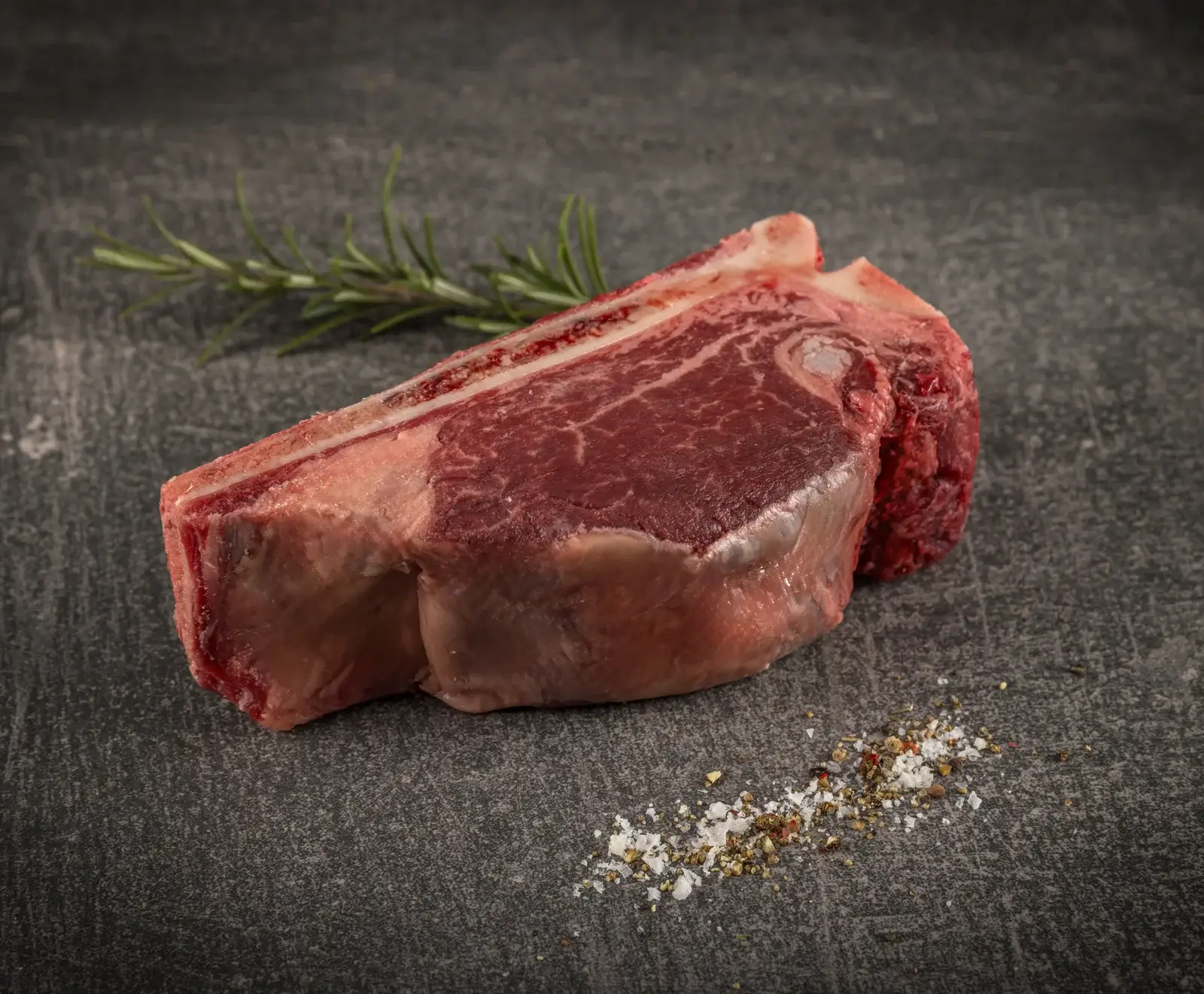 Ein rohes, marmoriertes Rindersteak mit Knochen auf dunklem Untergrund. Ein Rosmarinzweig liegt im Hintergrund, grobes Salz und Pfeffer im Vordergrund.