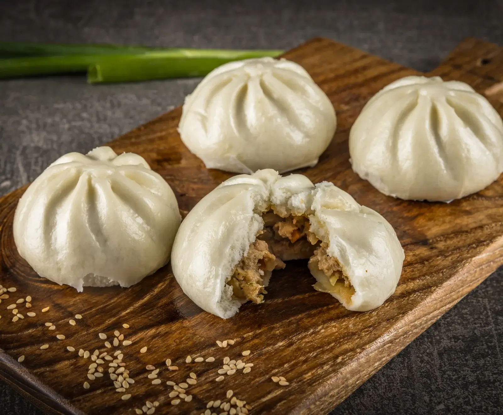 Vier gedämpfte Brötchen (Baozi) auf einem Holzbrett. Eines ist geöffnet und zeigt eine herzhafte Fleisch- und Gemüsefüllung. Sesamkörner sind verstreut, Frühlingszwiebeln im Hintergrund. Dunkler Untergrund.