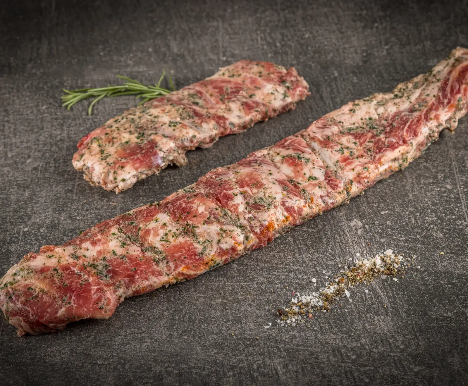 Zwei rohe Lammkoteletts mit Kräutern auf einer dunklen Schieferplatte, daneben etwas Salz.
