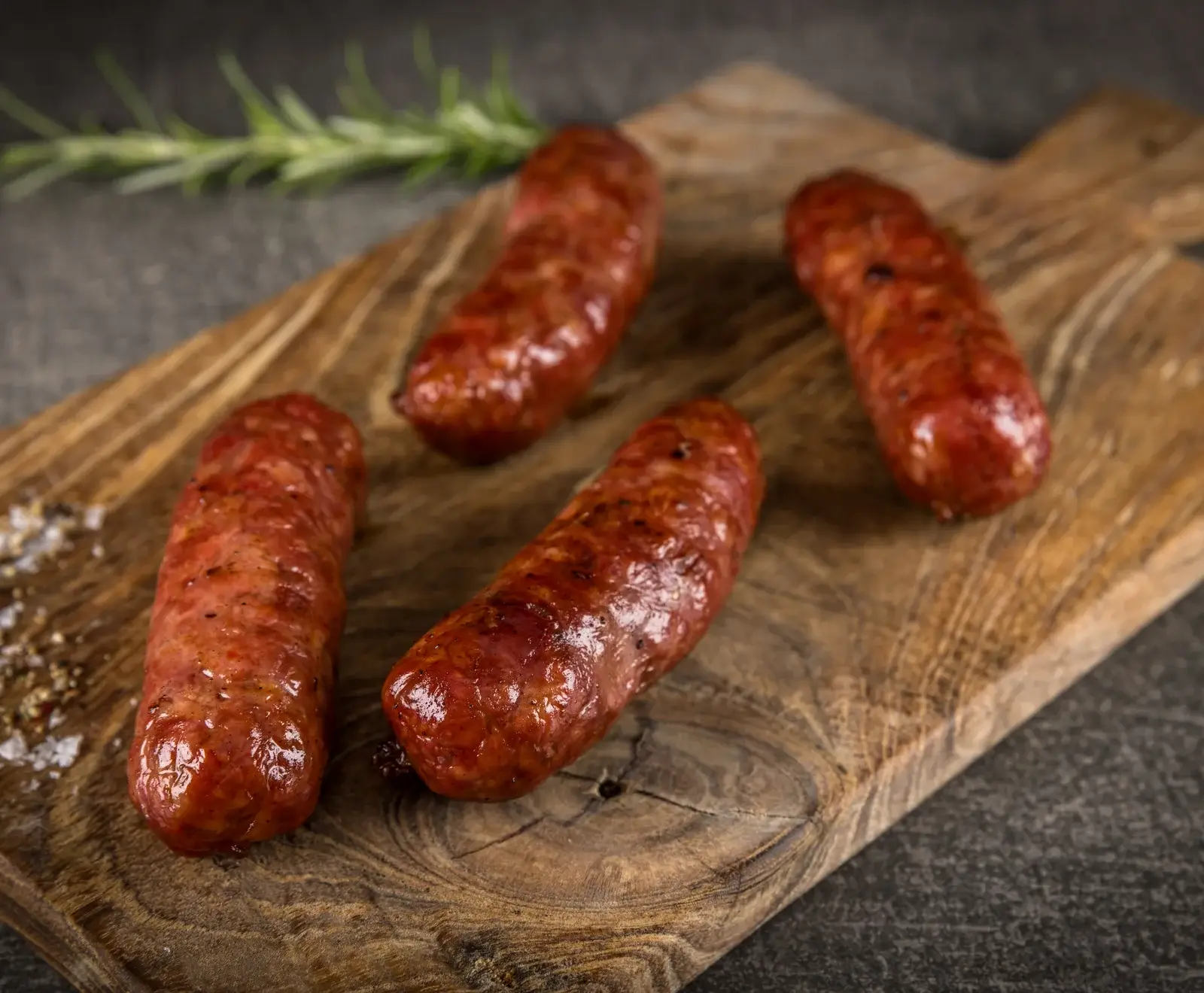Vier glänzende, gebratene Würste liegen auf einem rustikalen Holzbrett. Links daneben sind grobes Salz und Pfeffer, im Hintergrund ein Rosmarinzweig auf dunklem Untergrund zu sehen.