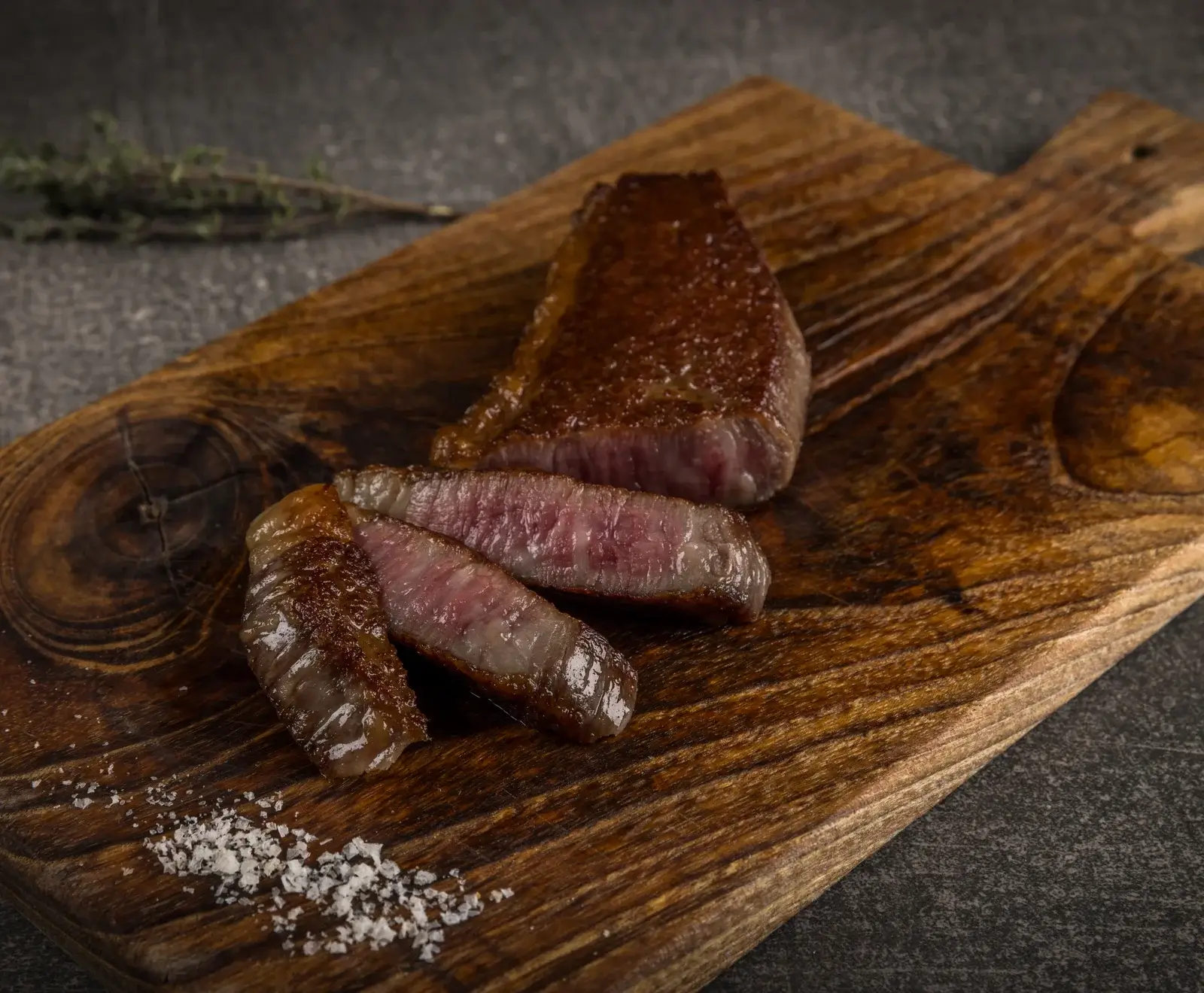 Ein perfekt gebratenes Steak, teilweise geschnitten, mit rosa Kern auf einem rustikalen Holzbrett. Daneben grobes Salz und ein Thymianzweig auf dunklem Hintergrund.