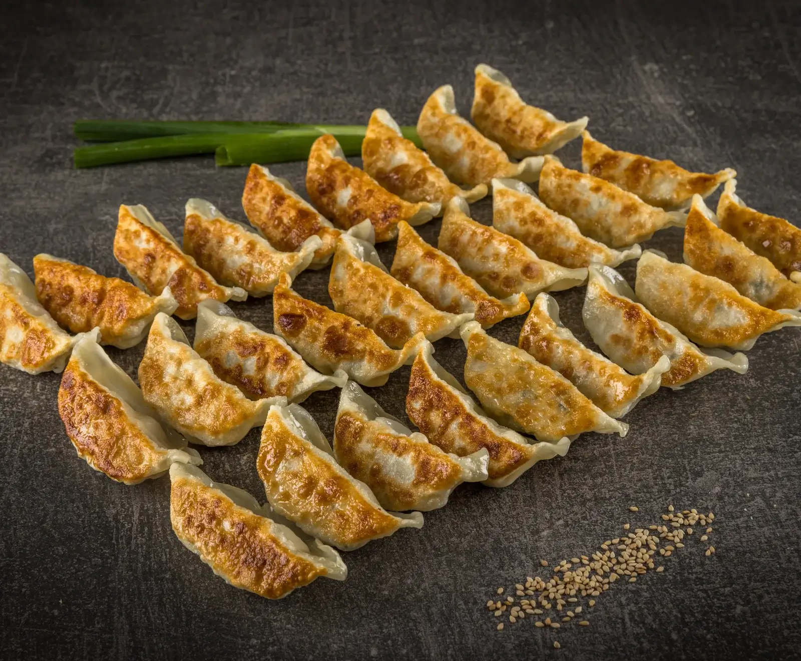 Nahaufnahme von zahlreichen goldbraun gebratenen Gyoza (Teigtaschen) in Reihen auf einer dunklen Oberfläche. Im Hintergrund liegen grüne Zwiebeln, im Vordergrund sind Sesamsamen verstreut.
