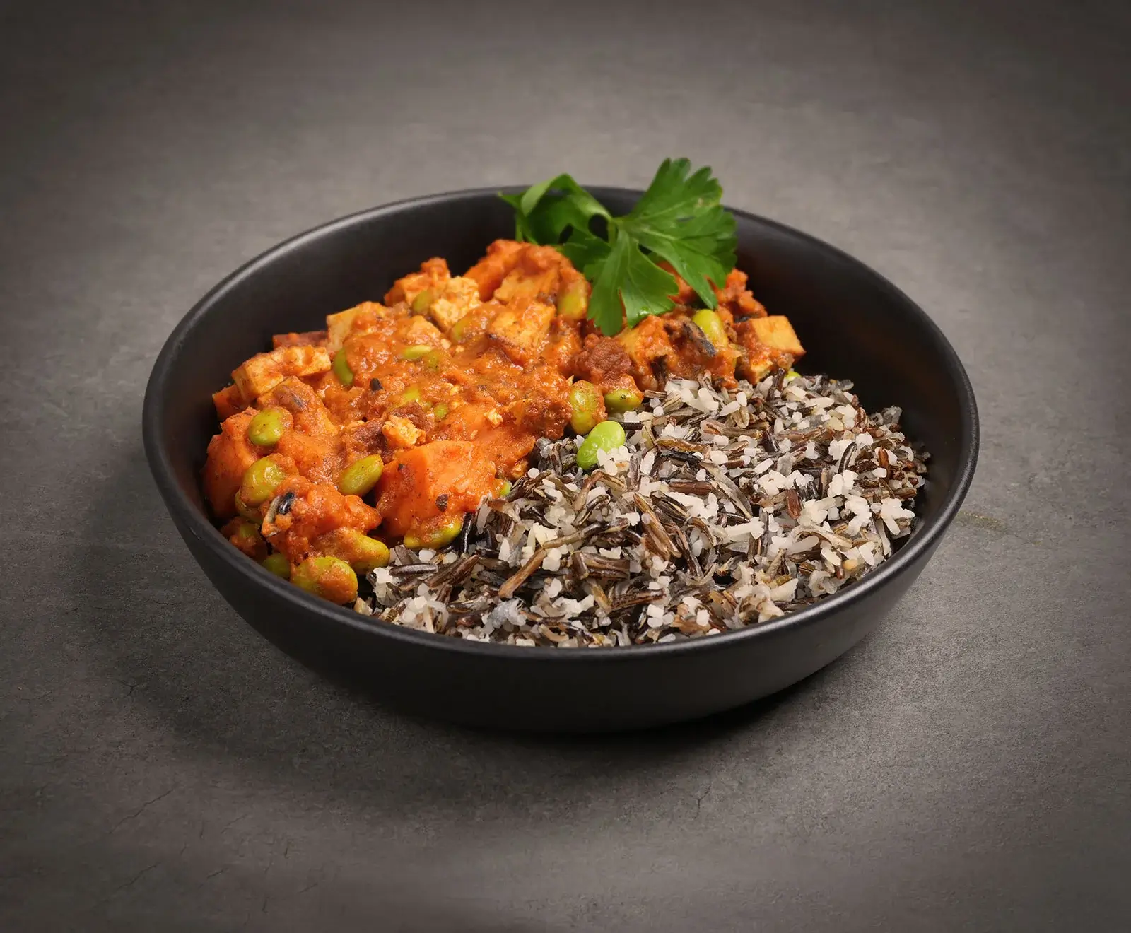 Schwarze Schale mit veganem Tofu-Curry, Edamame und Süßkartoffelstücken, serviert mit Wildreis und garniert mit Petersilie auf dunklem Hintergrund.