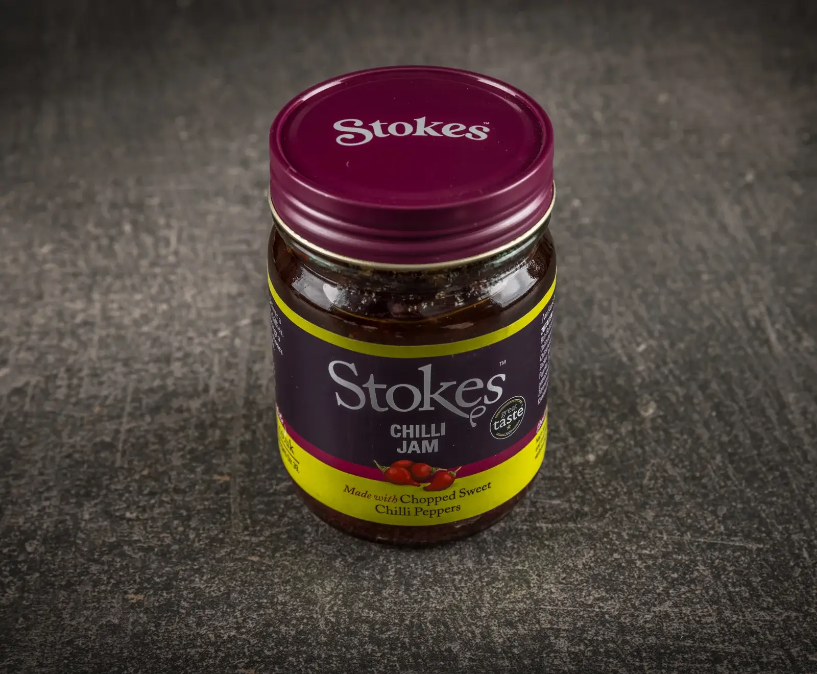 Ein Glas Stokes Chilli Jam mit dunkellila Deckel und lila-gelbem Etikett, das "Chilli Jam" und "great taste" zeigt, steht auf einer dunklen, strukturierten Oberfläche. Die Marmelade ist dunkelrotbraun mit Stücken.