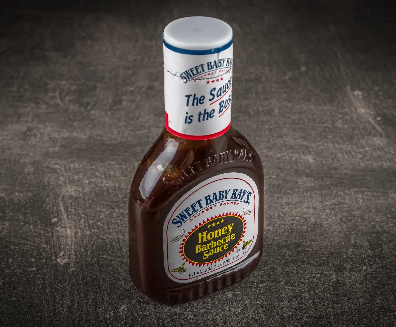 Eine Flasche Sweet Baby Ray's Honey Barbecue Sauce steht auf einer dunklen, strukturierten Oberfläche. Die braune Flasche hat ein weißes Etikett mit gelbem "Honey Barbecue Sauce"-Text und einem weißen Verschluss.
