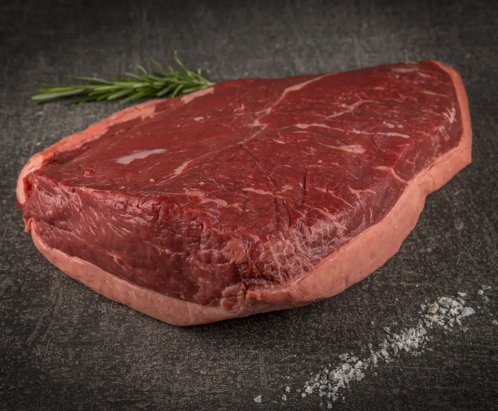 Ein großes, rohes Stück Rindfleisch, vermutlich Picanha, mit deutlichem Fettrand, liegt auf einer dunklen Steinplatte. Ein Rosmarinzweig und grobe Salzkristalle sind daneben platziert.