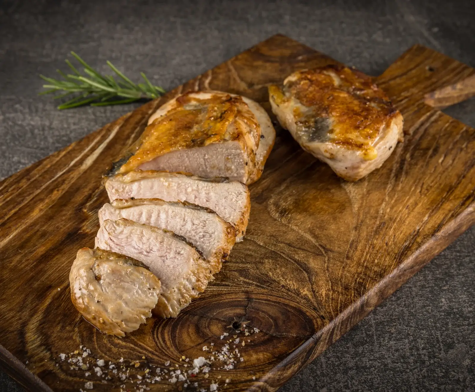 Zwei goldbraun gebratene Fleischstücke auf einem rustikalen Holzbrett. Eines ist in Scheiben geschnitten, daneben liegt ein ganzes Stück. Mit Rosmarin und grobem Salz garniert.