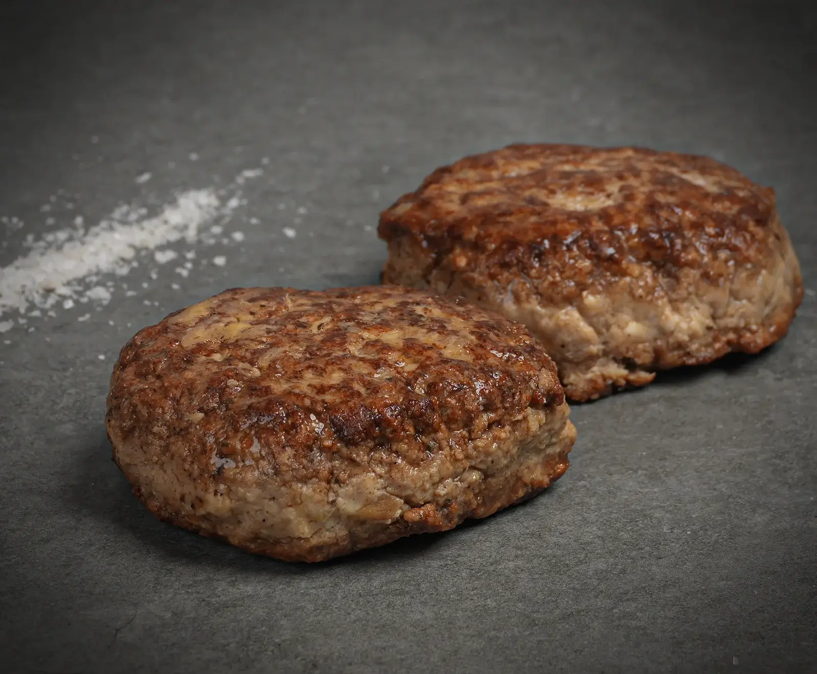 Zwei perfekt gebratene, runde Burger-Patties mit goldbrauner Kruste liegen auf einer dunklen Steinplatte. Links daneben ist eine Prise grobes Salz zu sehen.