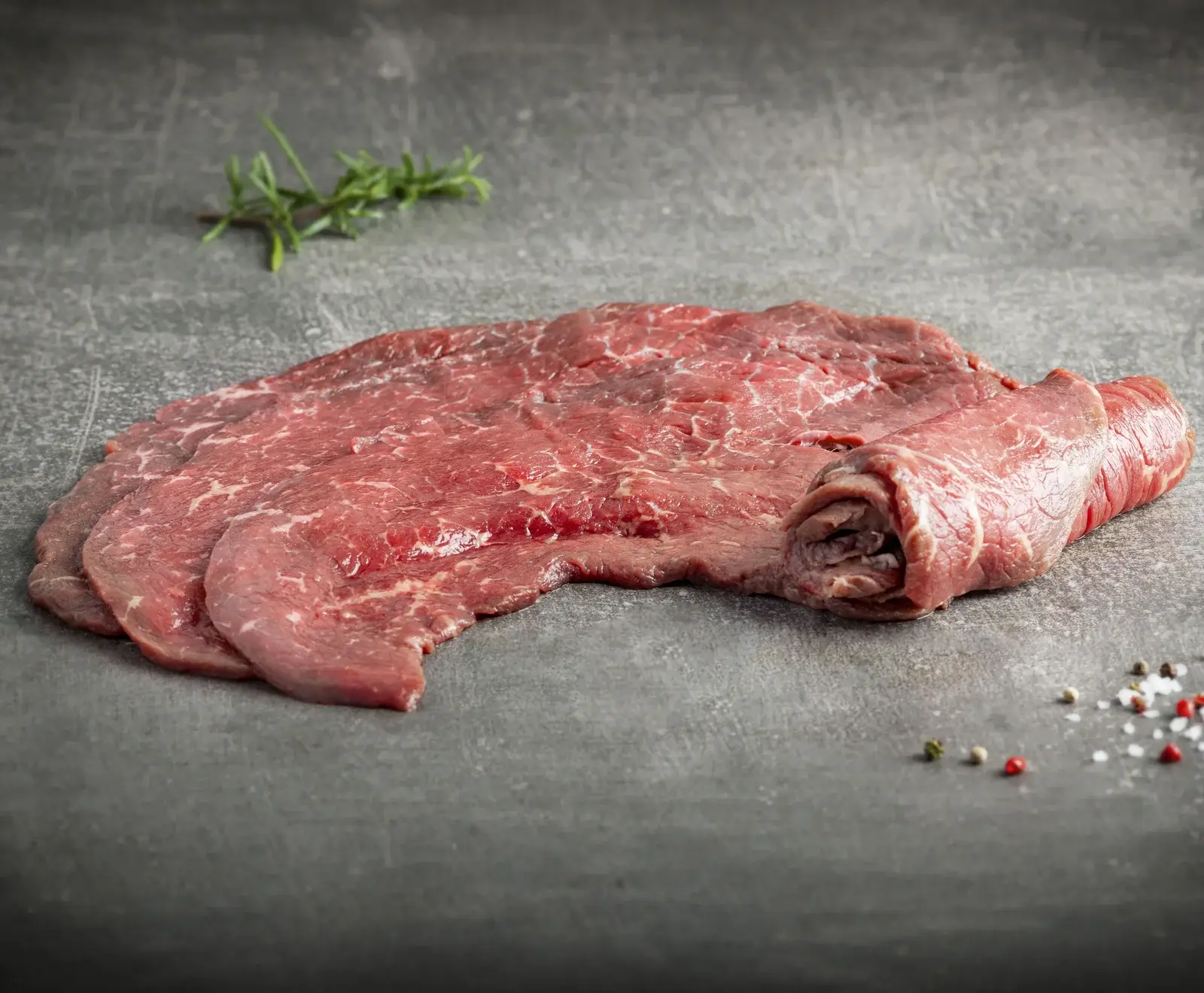Dünn geschnittenes, rohes Rindfleisch oder Kalbfleisch liegt auf einer dunklen, strukturierten Oberfläche. Mehrere Scheiben sind ausgebreitet, eine ist teilweise aufgerollt. Im Hintergrund ein Rosmarinzweig, im Vordergrund Pfefferkörner und grobes Salz