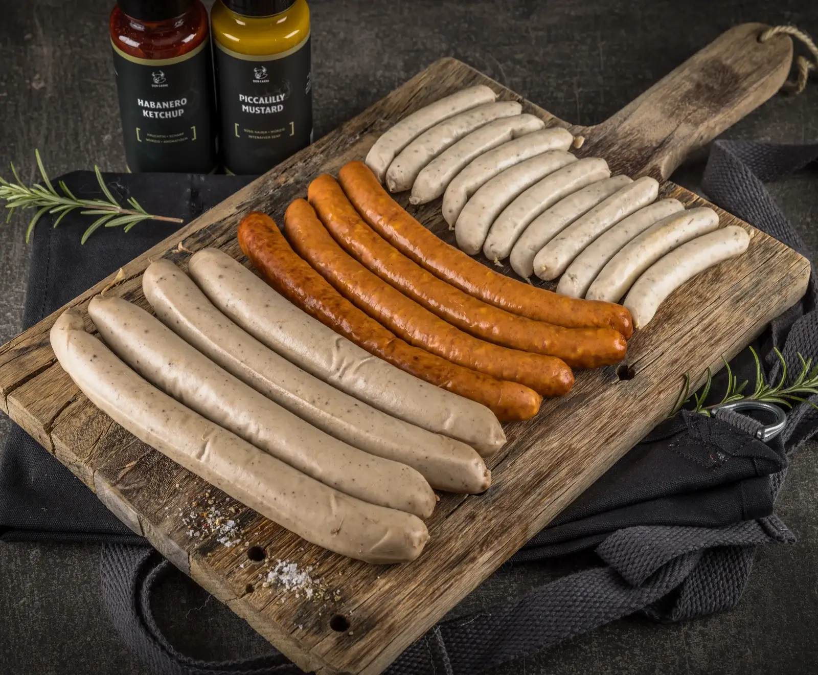 Bratwürste und Weißwürste auf einem Holzbrett, neben zwei Flaschen Senf und Rosmarin.