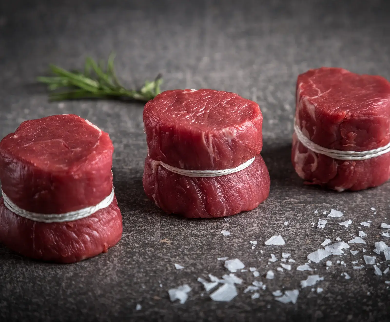 Drei rohe Rinderfiletsteaks, mit weißem Garn gebunden, liegen auf dunklem Untergrund. Eines ist doppelt gestapelt. Im Hintergrund Rosmarin, im Vordergrund grobes Meersalz.