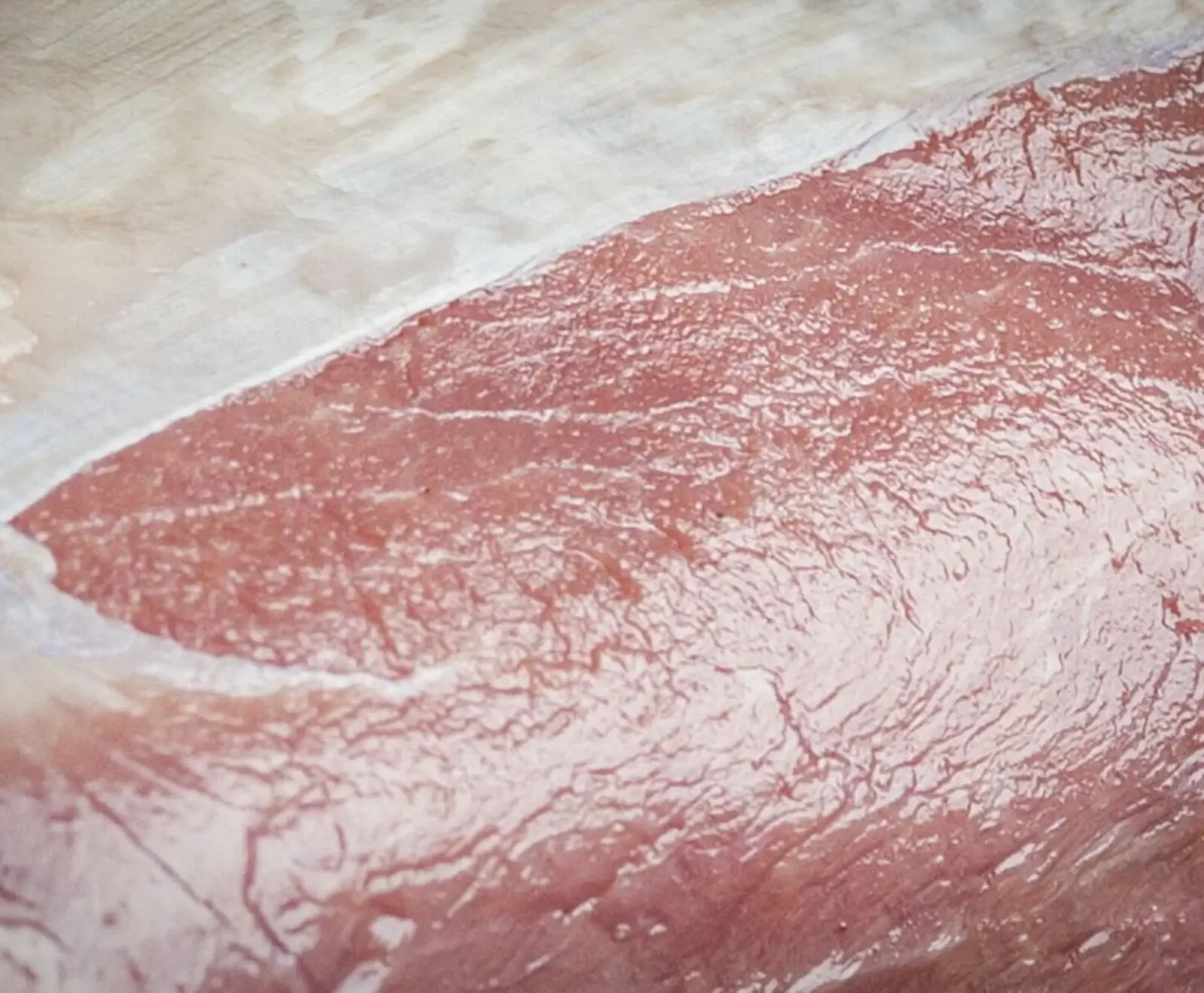 Nahaufnahme eines Stücks rohen, rötlich-rosa Fleisches mit sichtbaren Muskelfasern und einer dünnen Schicht weißem Fett oder Bindegewebe am oberen Rand.