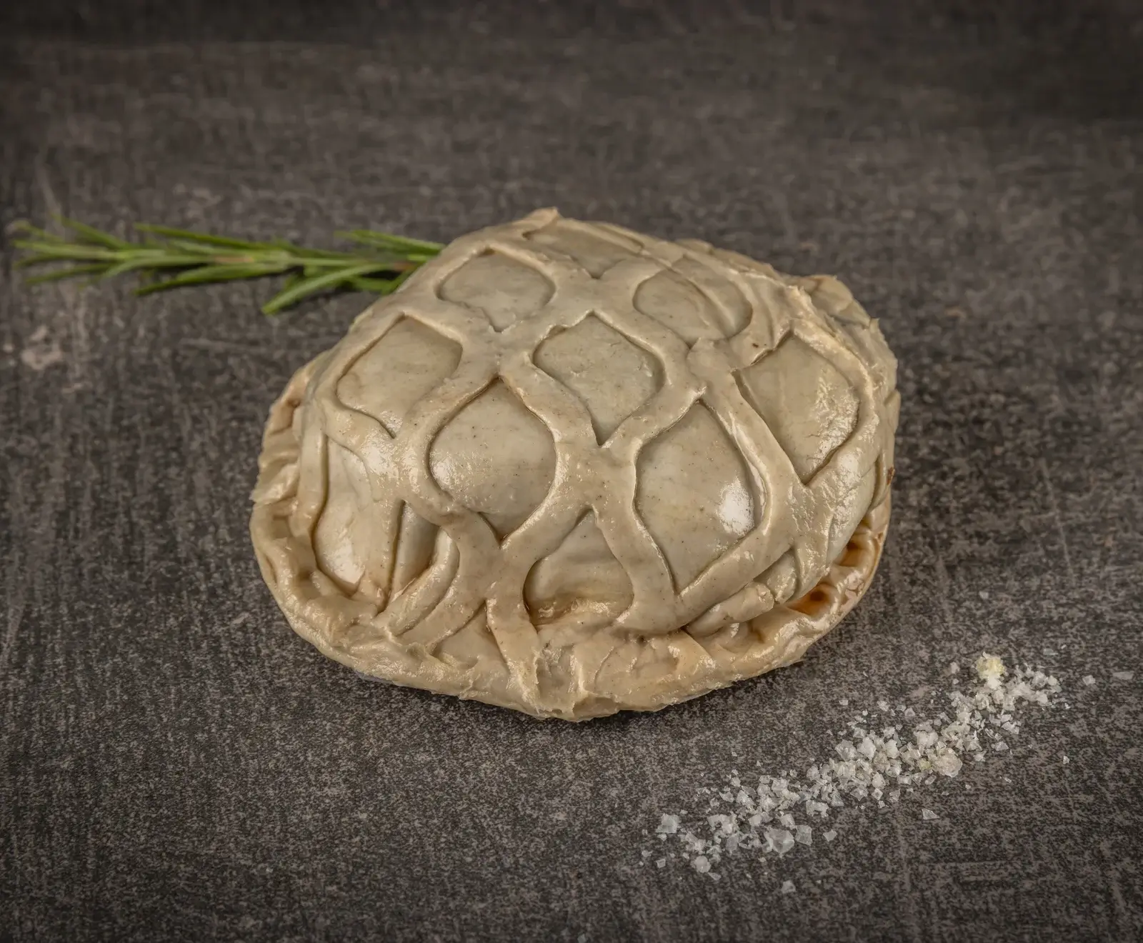 Rohes, hellbeiges Teiggebäck mit kunstvollem Gittermuster auf der Oberfläche, auf dunklem, strukturiertem Untergrund. Ein Rosmarinzweig und grobes Salz sind daneben platziert.