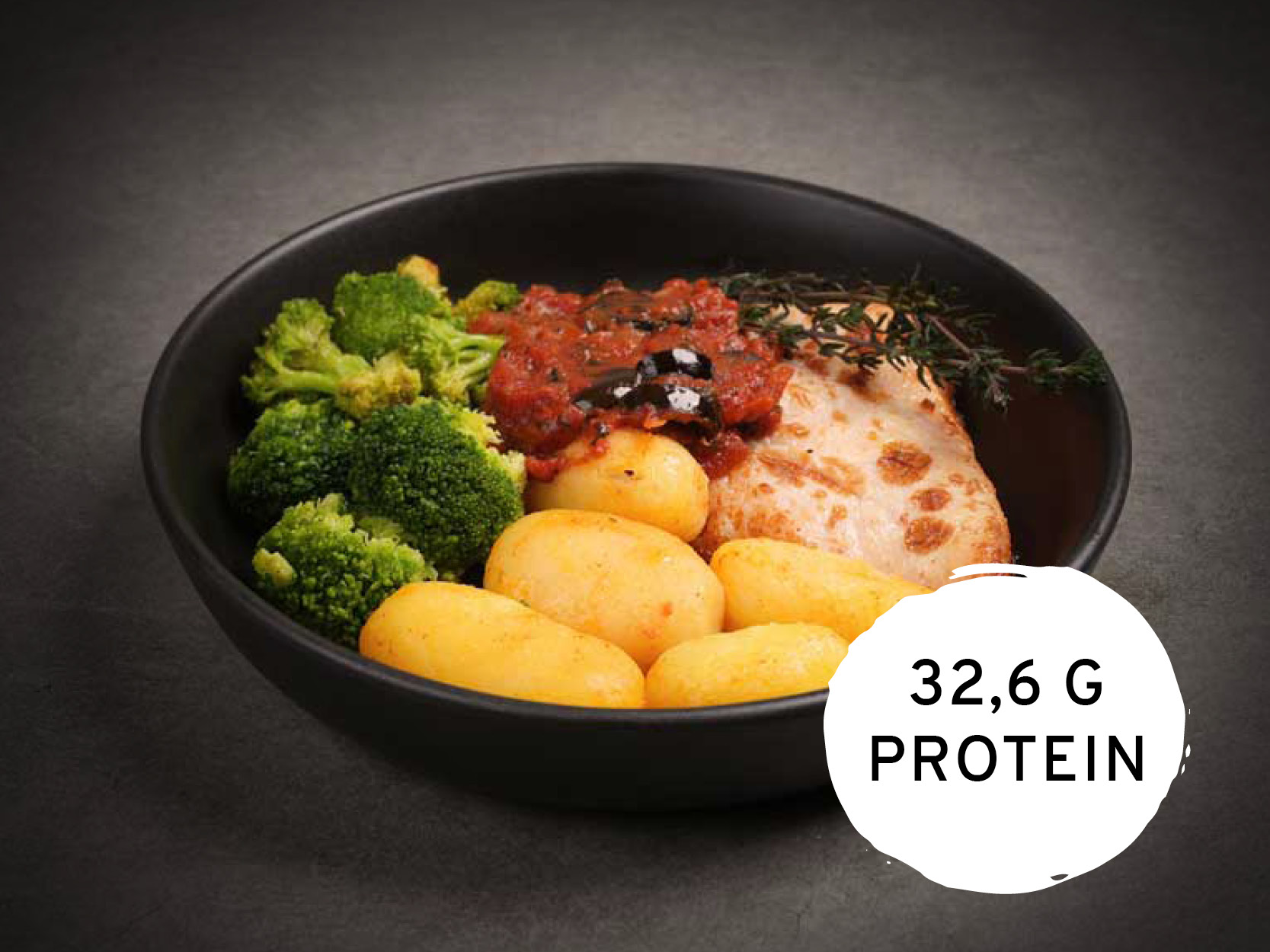 Eine Mahlzeit in einer schwarzen Schale mit Hähnchenbrust, Tomatensauce, kleinen Kartoffeln und Brokkoli, garniert mit Thymian. Ein weißer Kreis zeigt "32,6 G PROTEIN" an.