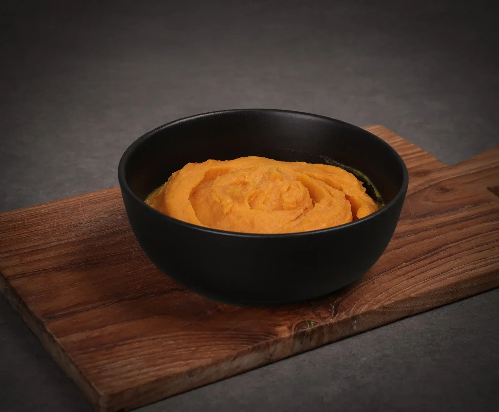 Eine schwarze Schale gefüllt mit leuchtend orangefarbenem, cremigem Püree, vermutlich Süßkartoffel- oder Kürbispüree, steht auf einem rustikalen Holzbrett vor dunklem Hintergrund. Die Oberfläche des Pürees ist leicht gewirbelt.