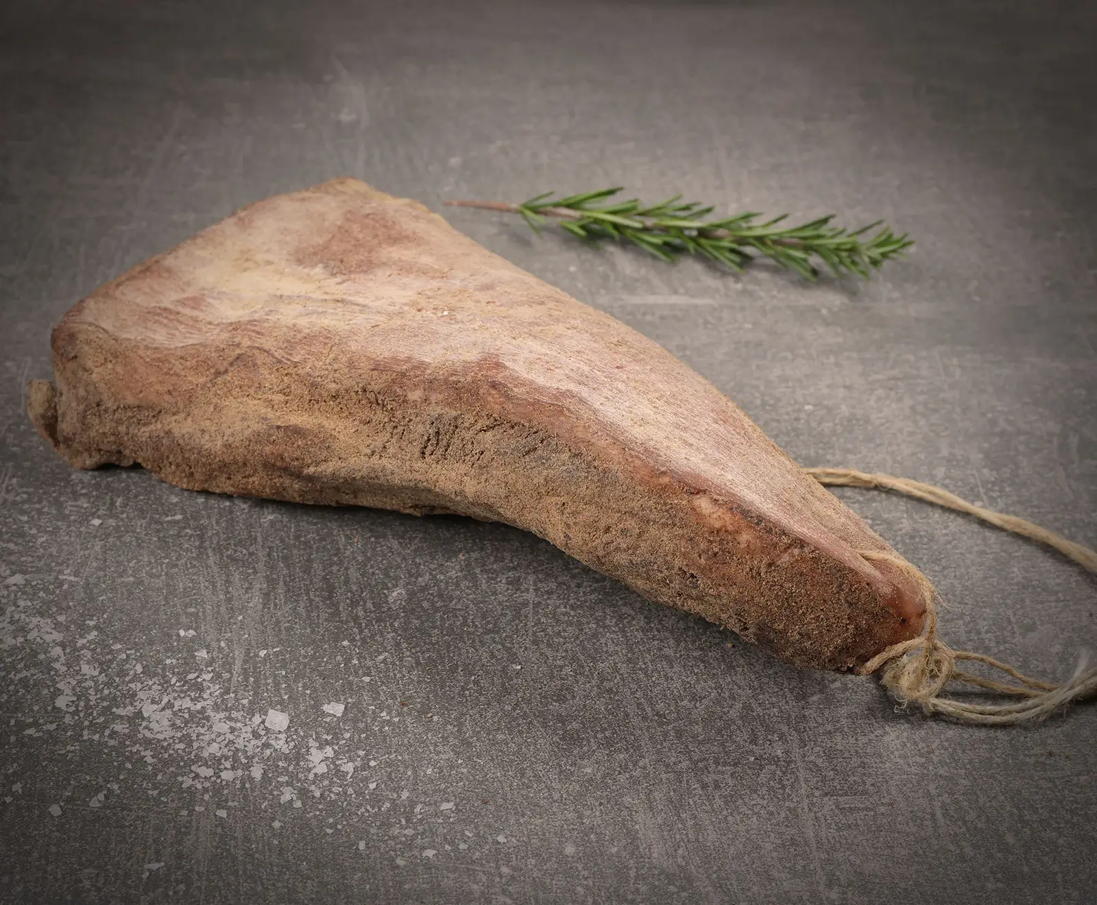 Ein keilförmiges Stück getrocknetes Fleisch mit rauer, bräunlicher Oberfläche, an einem Ende mit einer Schnur gebunden, liegt auf einem dunklen Untergrund mit Salzflocken. Im Hintergrund ein Rosmarinzweig.