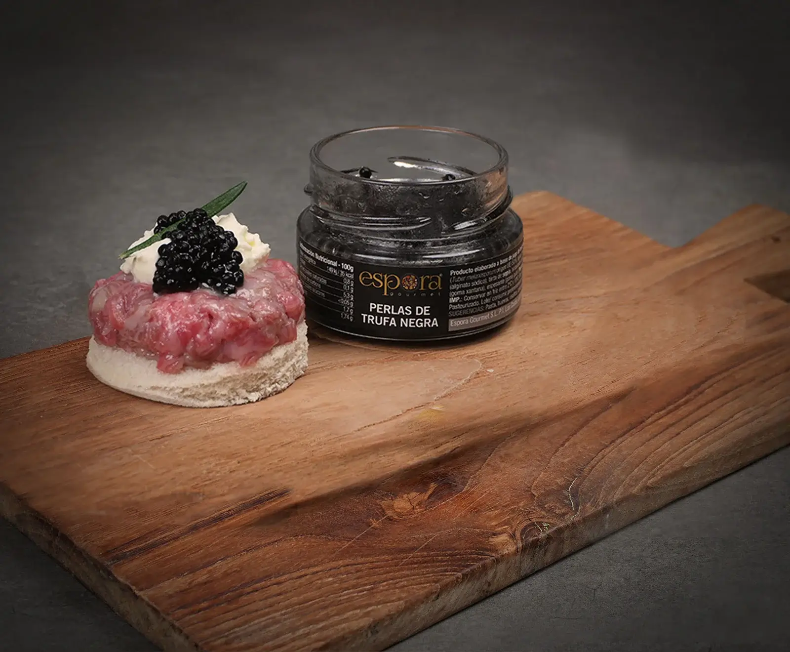 Ein kleines Glas "espora PERLAS DE TRUFA NEGRA" steht neben einem Canapé aus Steak-Tatar auf Weißbrot, garniert mit weißer Creme, schwarzen Trüffelperlen und einem grünen Blatt, alles auf einem Holzbrett.