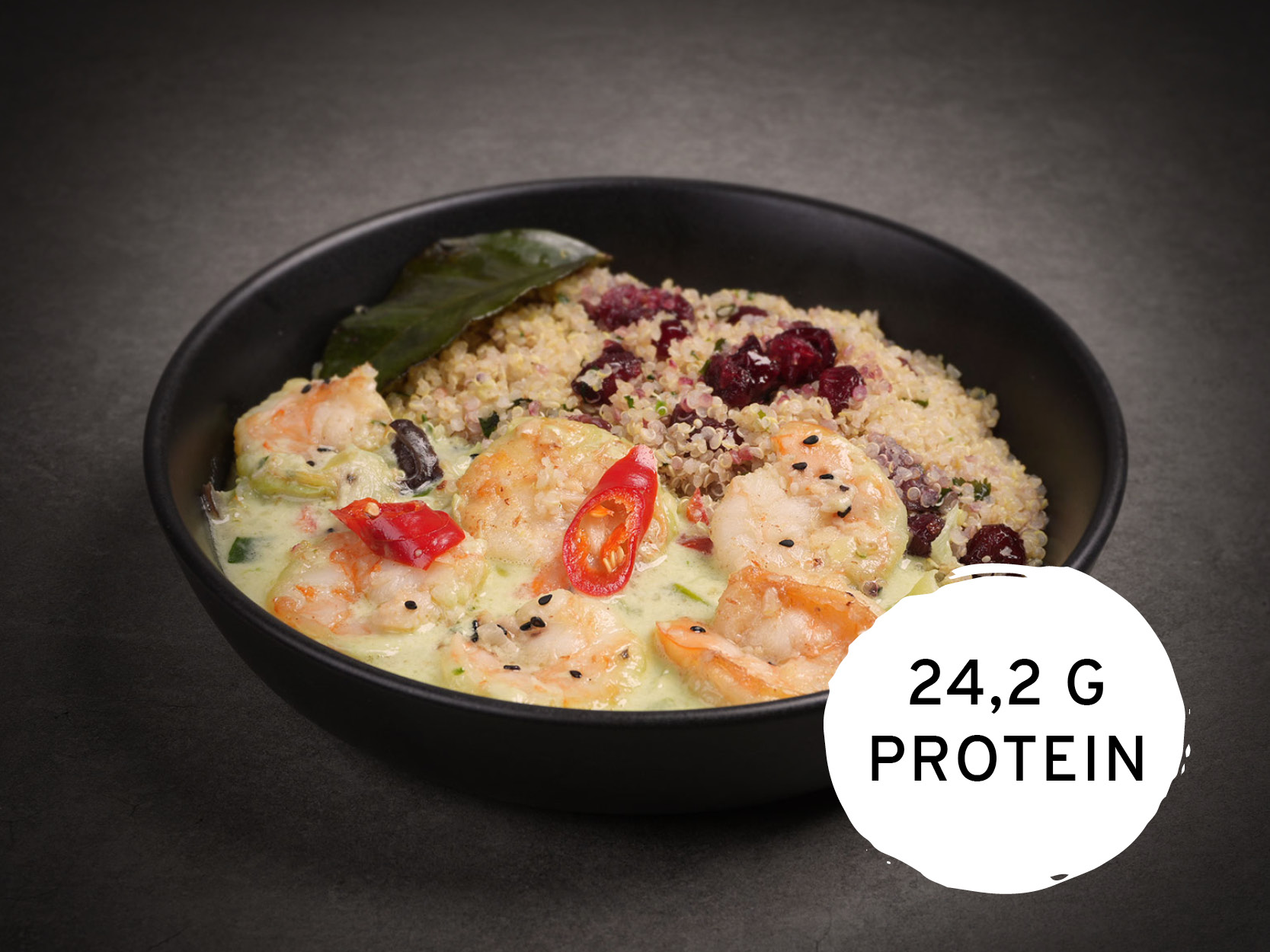 Eine schwarze Schale mit Garnelen in grüner Currysauce, Quinoa mit Cranberries, garniert mit Chilischeiben, schwarzem Sesam und einem Limettenblatt. Unten rechts steht "24,2 G PROTEIN".