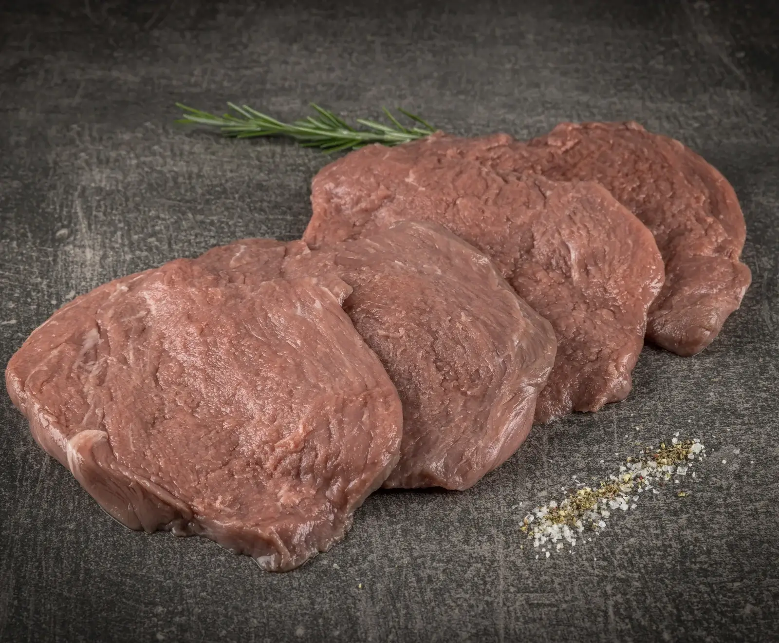 Vier rohe, dünne Fleischscheiben liegen diagonal auf einer dunklen Steinplatte. Im Hintergrund ist ein Rosmarinzweig zu sehen, im Vordergrund eine Prise Salz und Pfeffer.