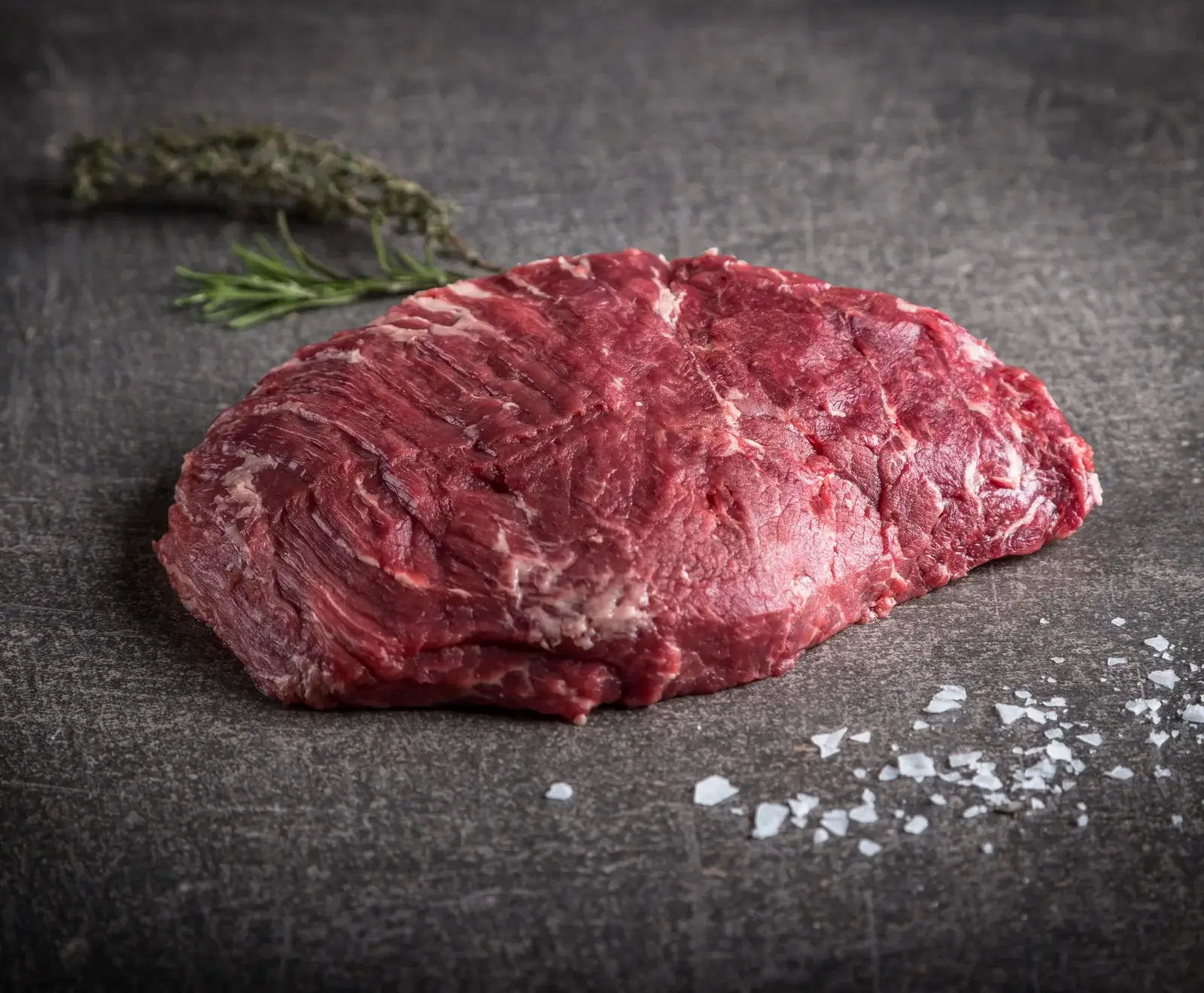 Ein rohes, marmoriertes Rindersteak liegt auf einer dunklen Oberfläche, umgeben von frischem Rosmarin, Thymian und groben Meersalzflocken.