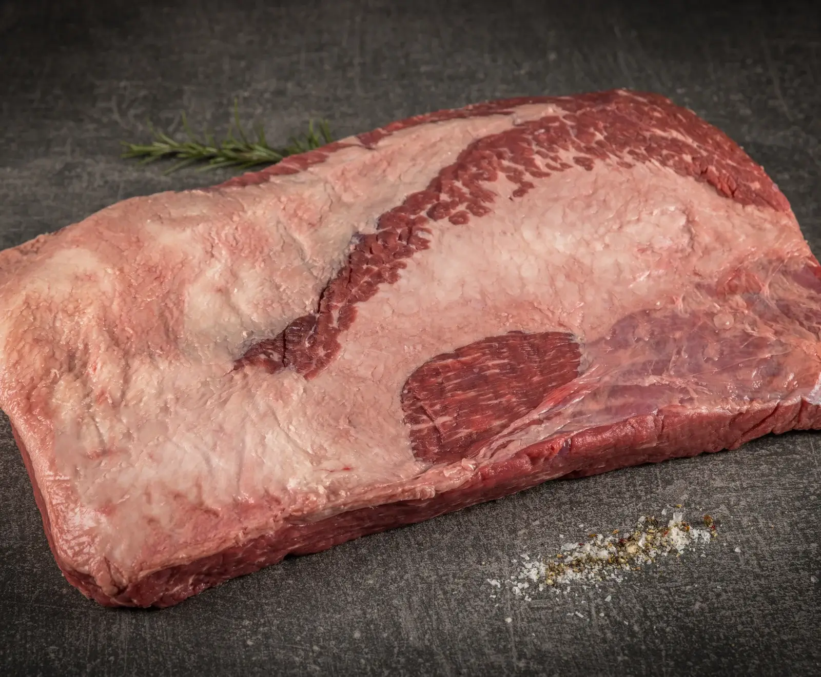 Rohes Rinderbruststück (Brisket) mit Fettschicht auf dunklem Untergrund. Ein Rosmarinzweig und eine Prise Salz und Pfeffer liegen daneben.
