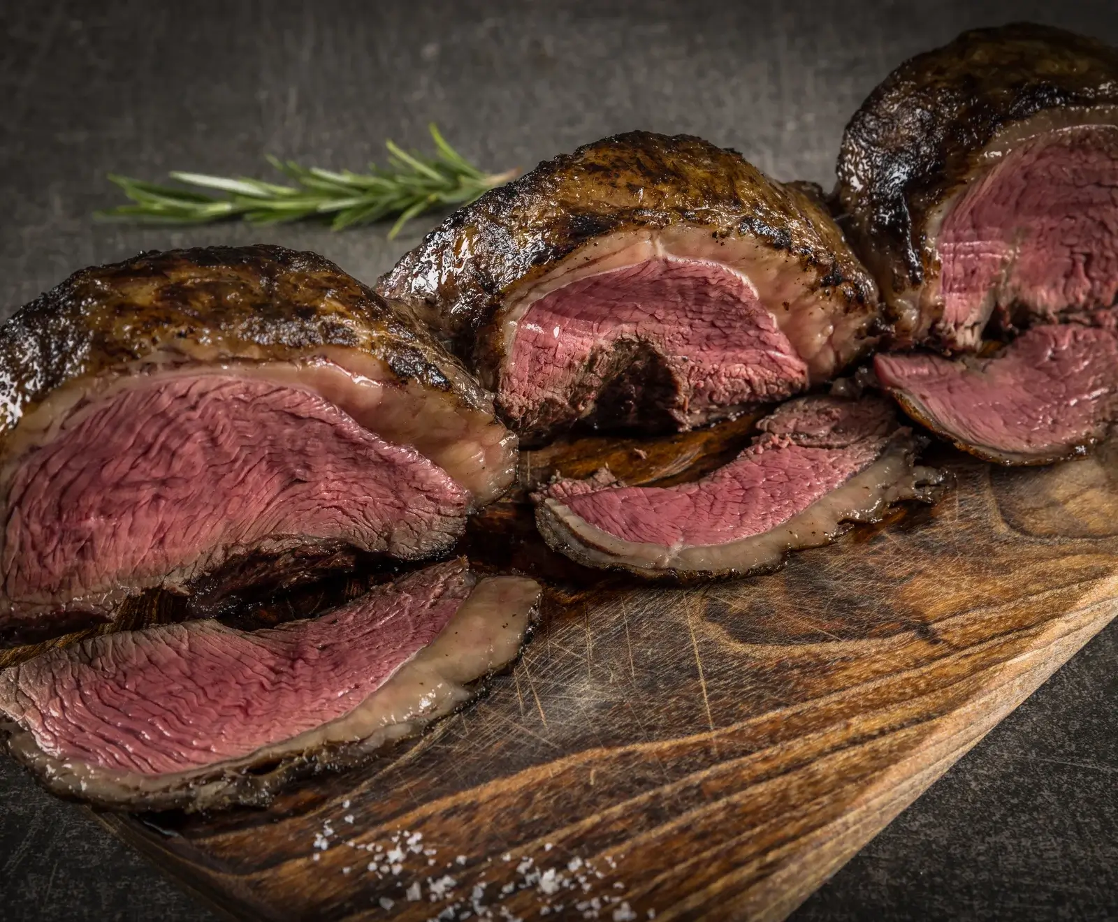 Nahaufnahme von aufgeschnittenem, medium-rare gegartem Picanha-Rindfleisch mit knuspriger Kruste auf einem Holzbrett, garniert mit Rosmarin und Meersalz.