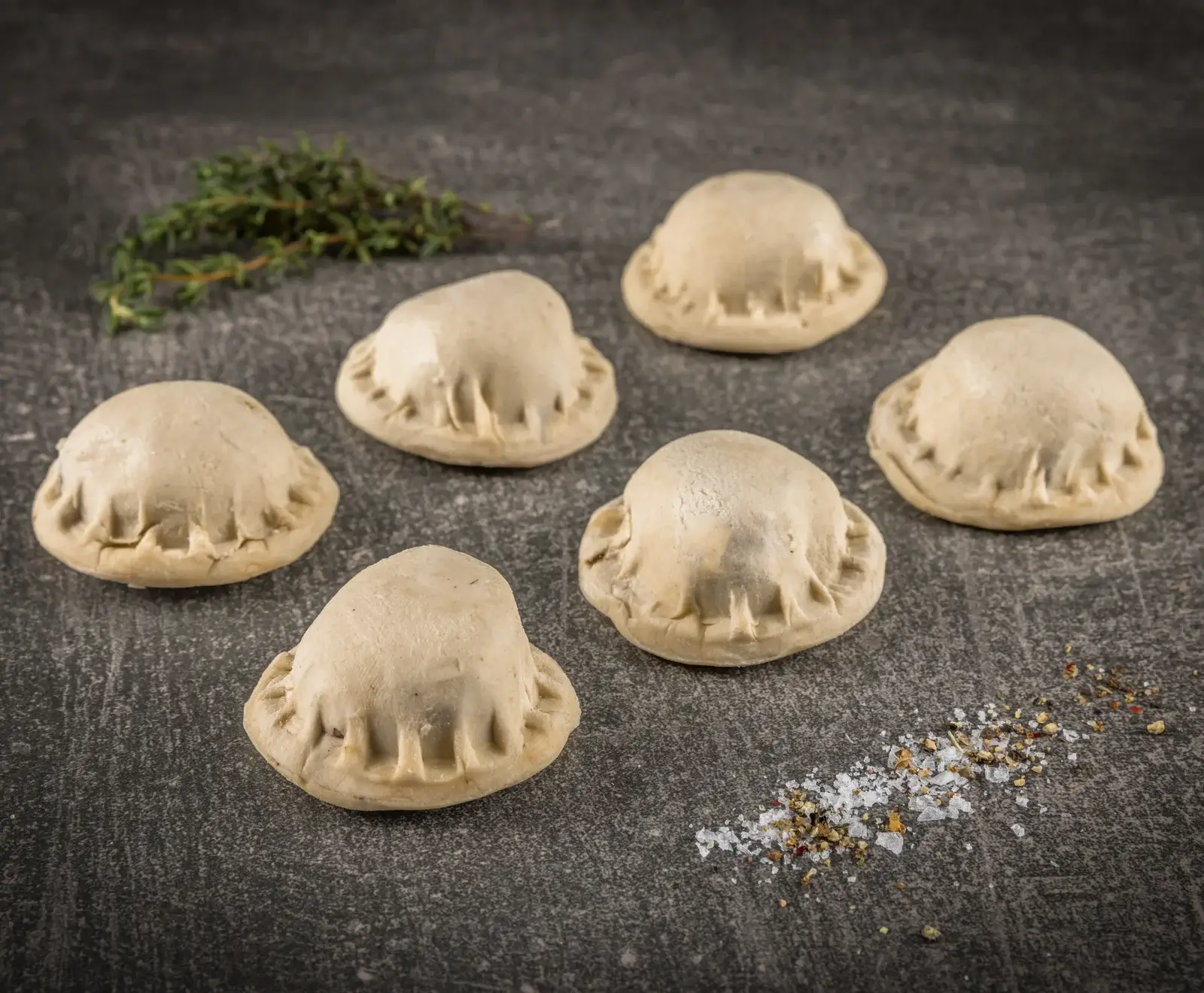 Sechs ungekochte, helle Teigtaschen mit gewelltem Rand auf dunklem Untergrund, daneben frischer Thymian, grobes Salz und Pfeffer.