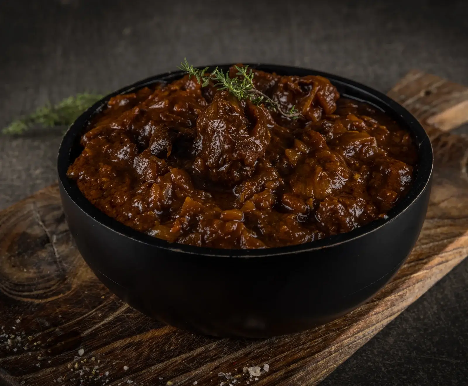 Eine schwarze Schale gefüllt mit dunklem, reichhaltigem Gulasch oder Eintopf, garniert mit einem Thymianzweig, auf einem rustikalen Holzbrett mit verstreutem Salz und Pfeffer.