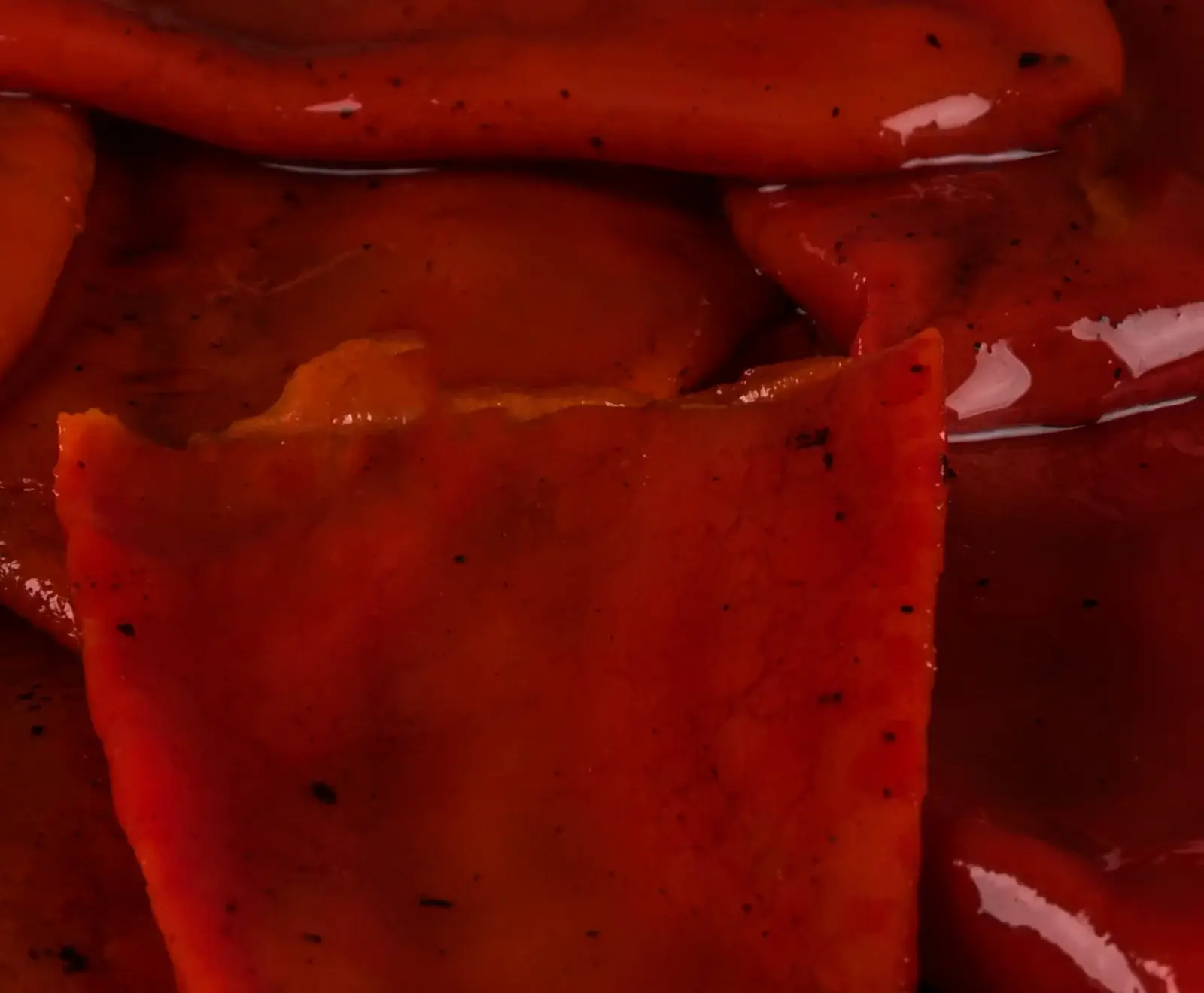 Nahaufnahme von mehreren glänzenden Stücken gerösteter roter Paprika, die in einer Flüssigkeit liegen. Die Paprika zeigt eine tiefrote Farbe mit leichten Bräunungsspuren und dunklen Gewürzpartikeln.