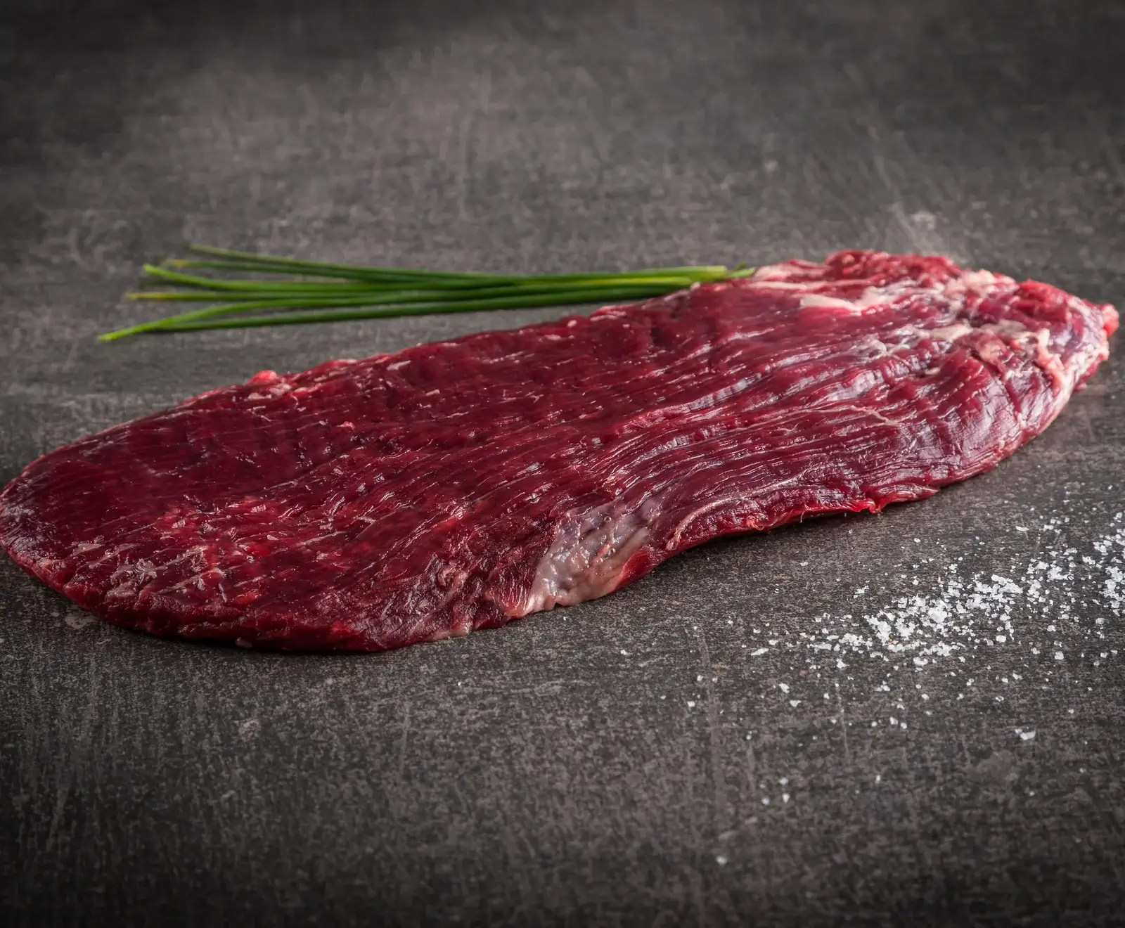 Ein rohes, dunkelrotes Flank Steak liegt auf einer dunklen, strukturierten Oberfläche. Dahinter liegen grüne Schnittlauchhalme, rechts sind grobe Salzkristalle verstreut.