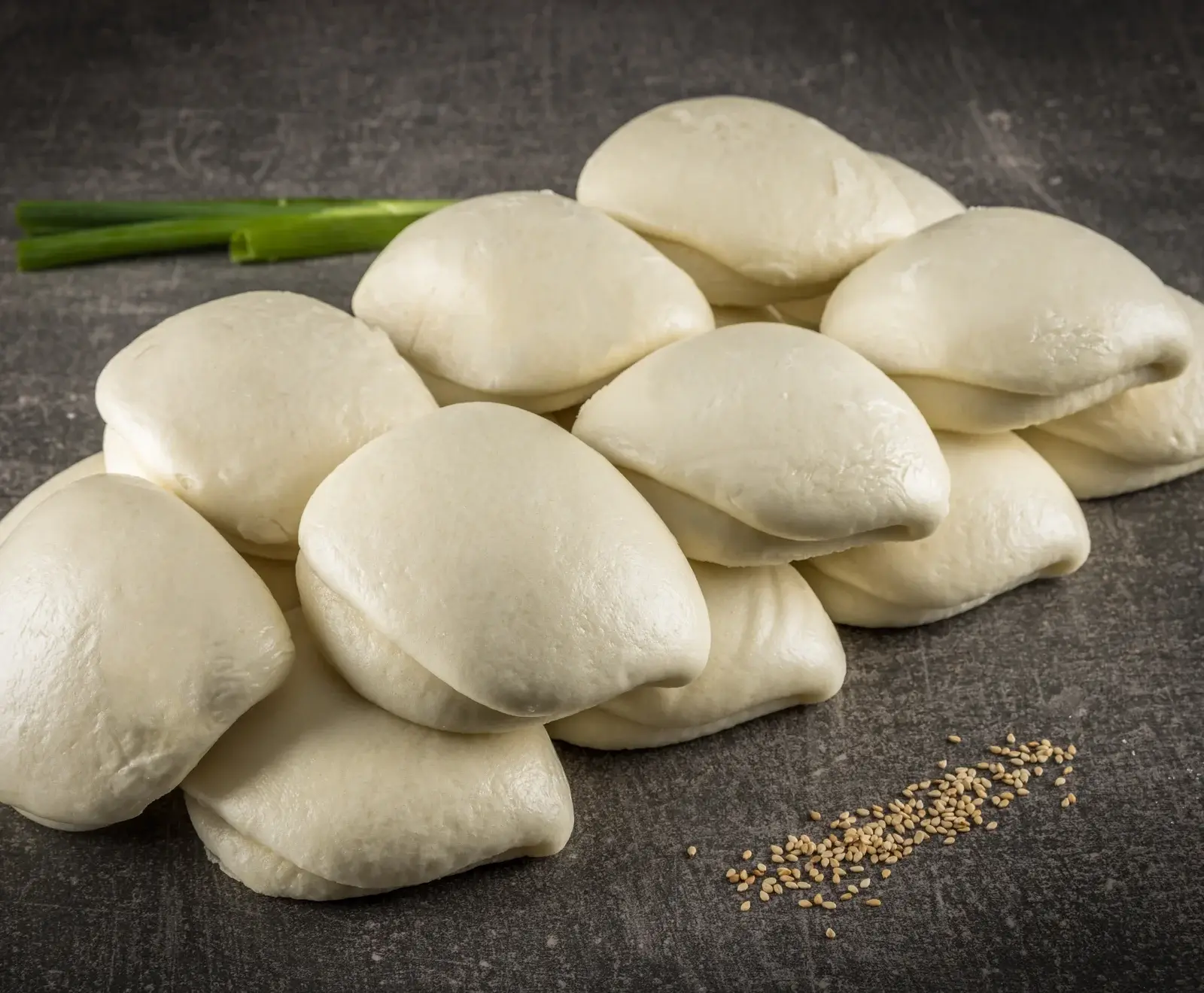Ein Stapel weicher, weißer gedämpfter Bao Buns liegt auf einer dunklen, strukturierten Oberfläche. Im Hintergrund sind grüne Lauchzwiebeln und im Vordergrund Sesamsamen zu sehen.