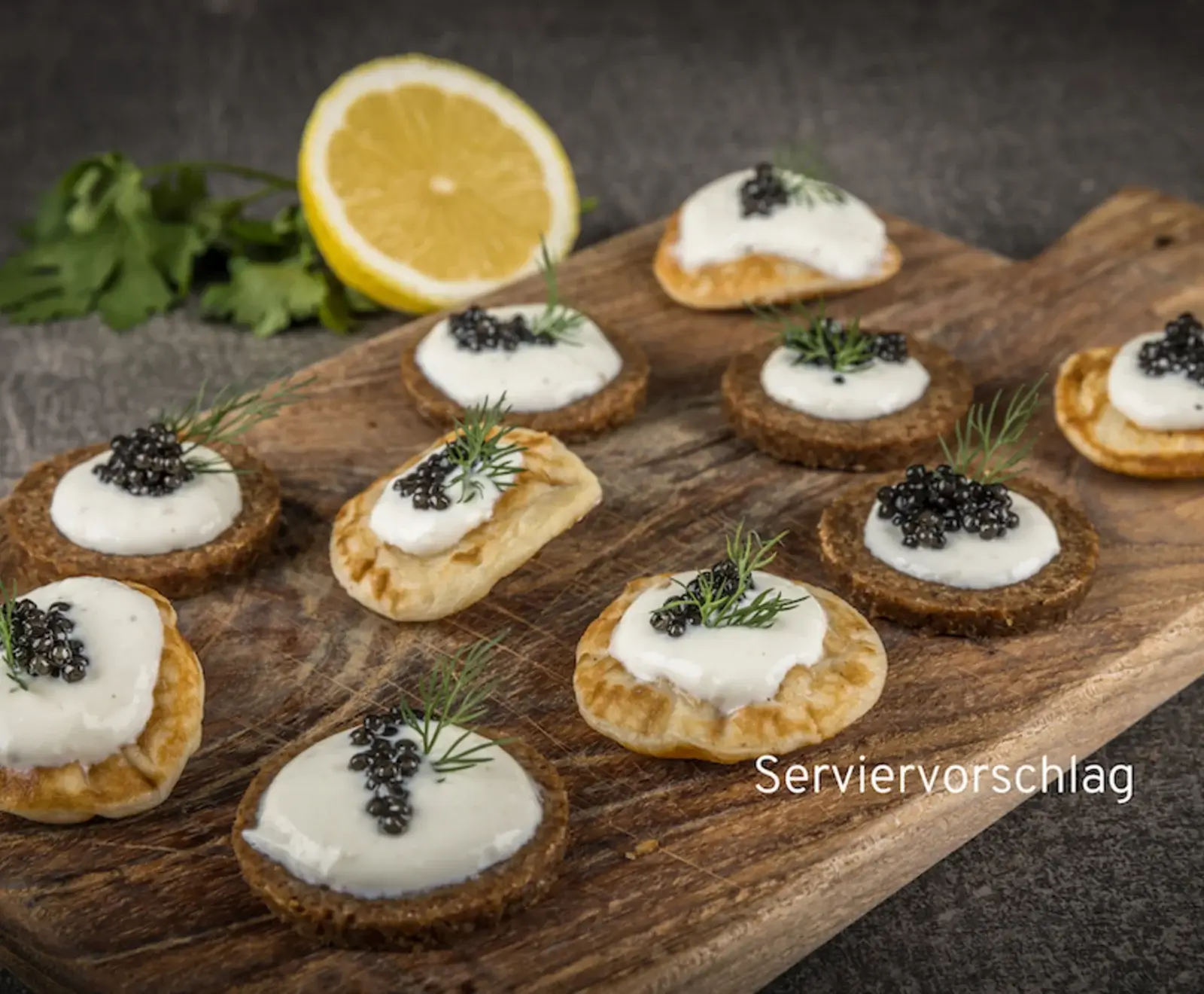 Nahaufnahme von mehreren Canapés mit schwarzem Kaviar, weißer Creme und Dill auf verschiedenen Brot- oder Gebäckbasen, angerichtet auf einem Holzbrett. Im Hintergrund eine halbe Zitrone.