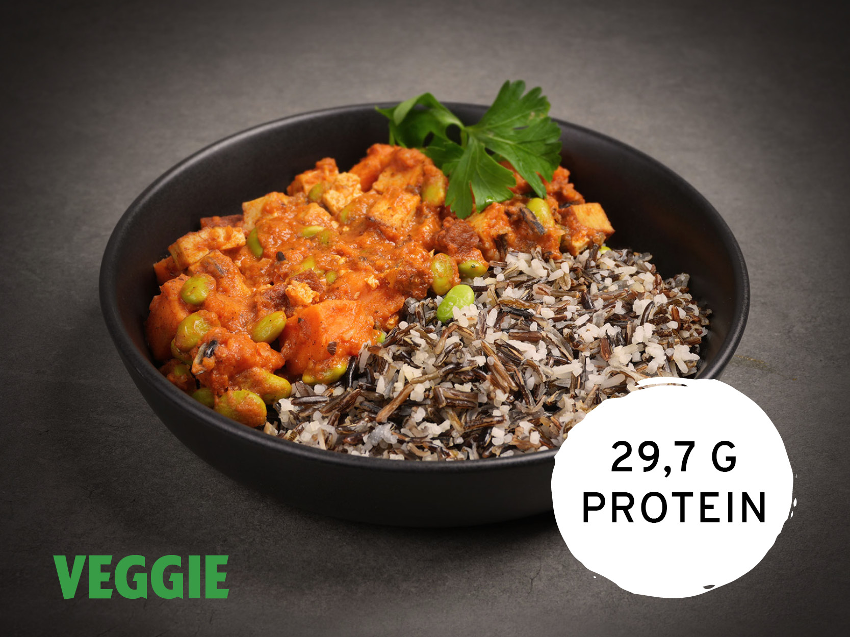 Eine schwarze Schale mit Tofu-Curry, Edamame und Wildreis, garniert mit Petersilie. Das Gericht ist als "VEGGIE" gekennzeichnet und enthält 29,7 g Protein.