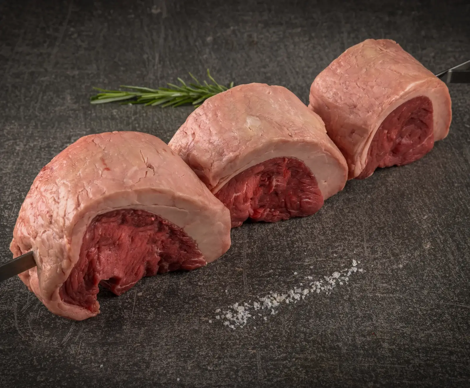 Drei rohe Picanha-Stücke (Tafelspitz mit Fettrand) auf einem Metallspieß, liegend auf einer dunklen Steinplatte. Daneben liegen ein Rosmarinzweig und grobes Meersalz.