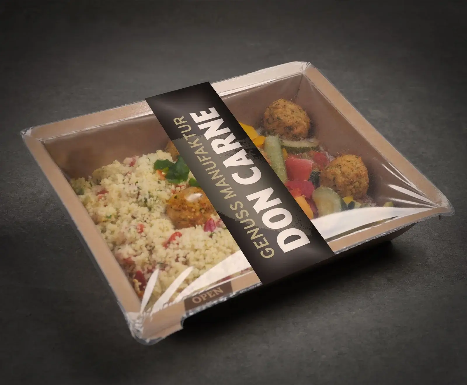 Eine rechteckige, braune Essensbox mit Couscous, Gemüse und Falafel, versiegelt mit Folie und einem schwarzen "DON CARNE" Etikett, auf dunklem Hintergrund.