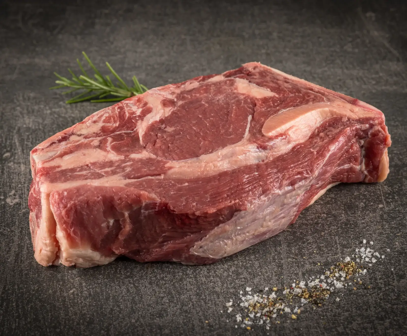 Ein rohes, marmoriertes Rindersteak mit Fettrand liegt auf dunklem Untergrund. Daneben sind ein Rosmarinzweig sowie gestreutes Salz und Pfeffer zu sehen.