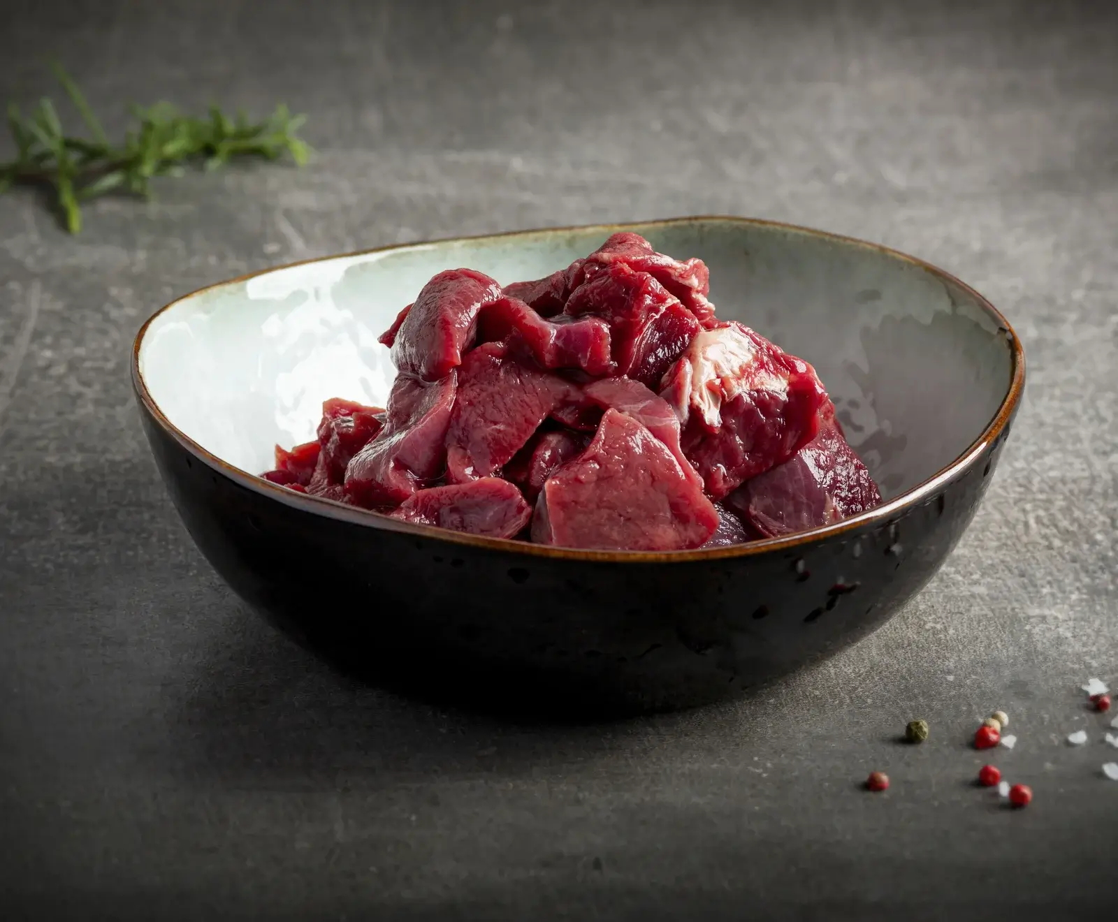 Eine dunkle Keramikschale gefüllt mit rohen, dunkelroten Fleischstücken (vermutlich Innereien wie Leber oder Herz) auf einem grauen Steinhintergrund. Im Vordergrund liegen Pfefferkörner und Salz, im Hintergrund Rosmarin.
