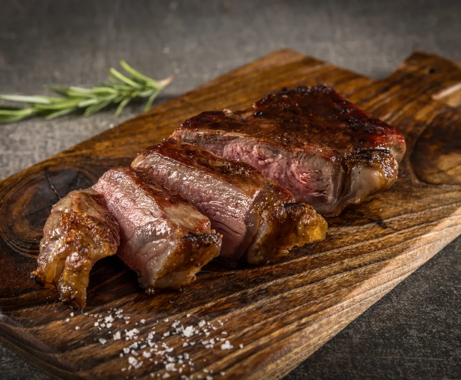 Ein perfekt gebratenes, in Scheiben geschnittenes Steak auf einem rustikalen Holzbrett, mit grobem Salz und einem Rosmarinzweig im Hintergrund.